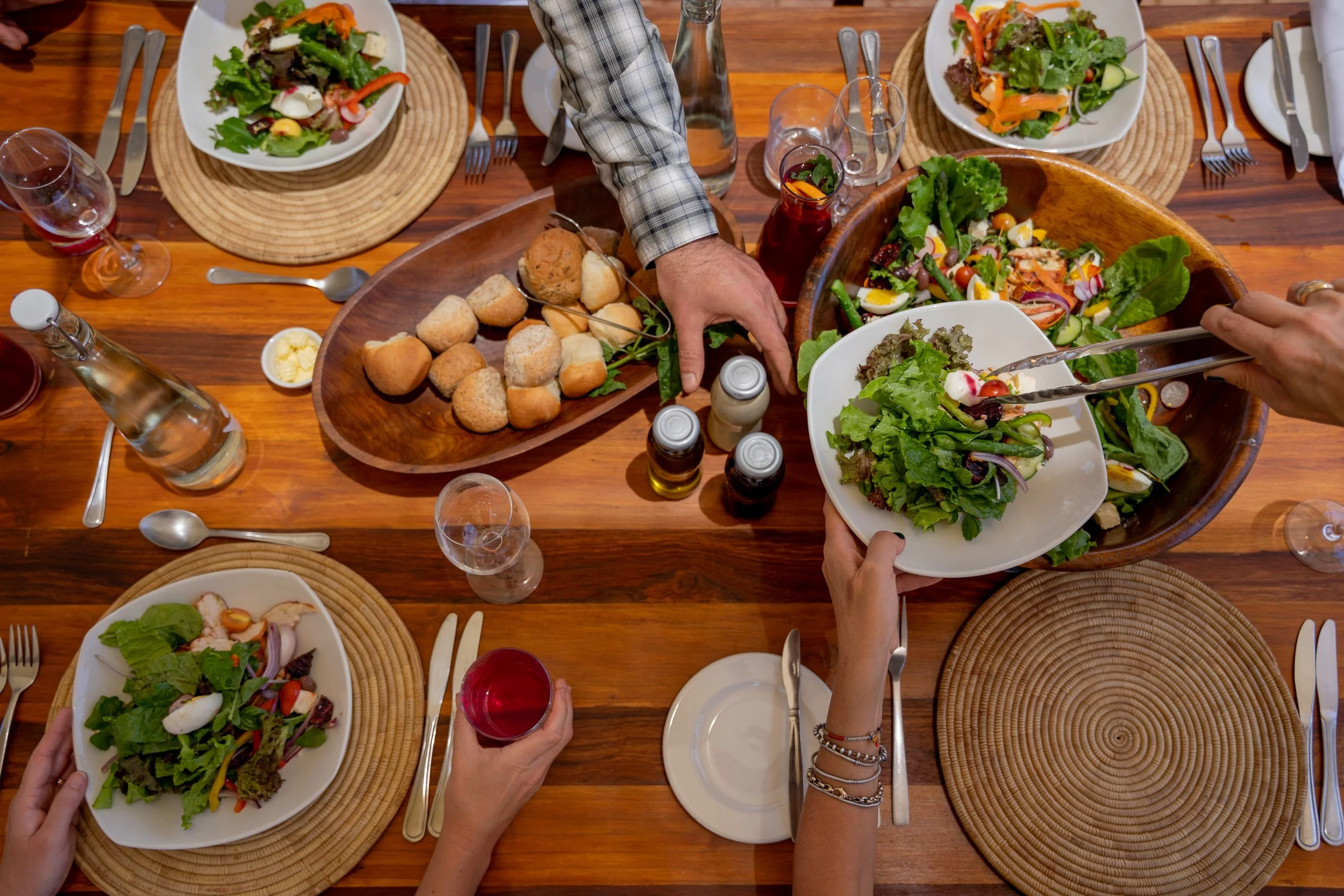 A top-down view of a dinner table with various salads, bread rolls, and drinks. Hands are seen reaching for salad and pouring drinks.