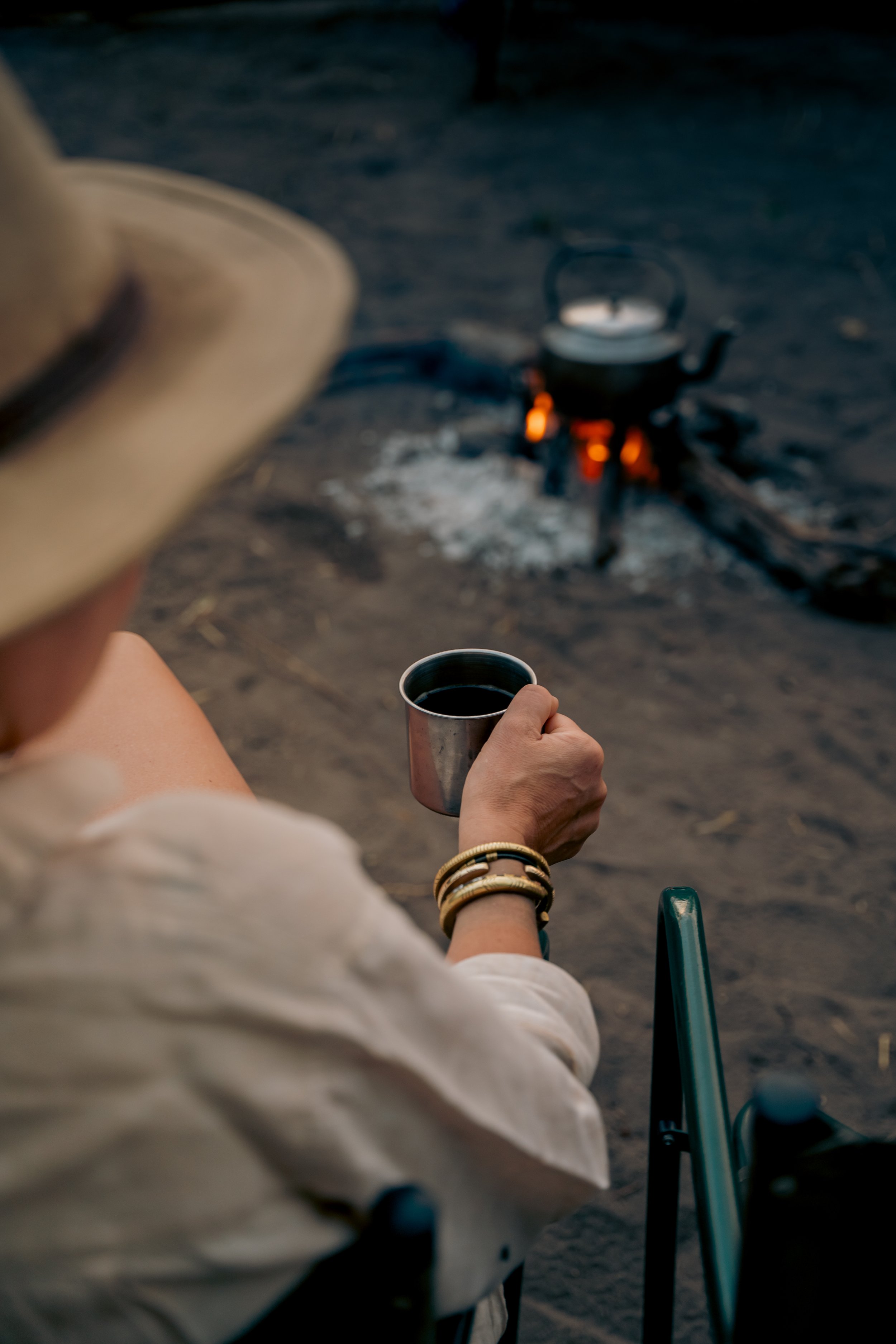Person in a wheelchair holding a metal mug with a dark beverage, sitting outdoors near a campfire with a kettle on it, in a natural setting during dusk or night.