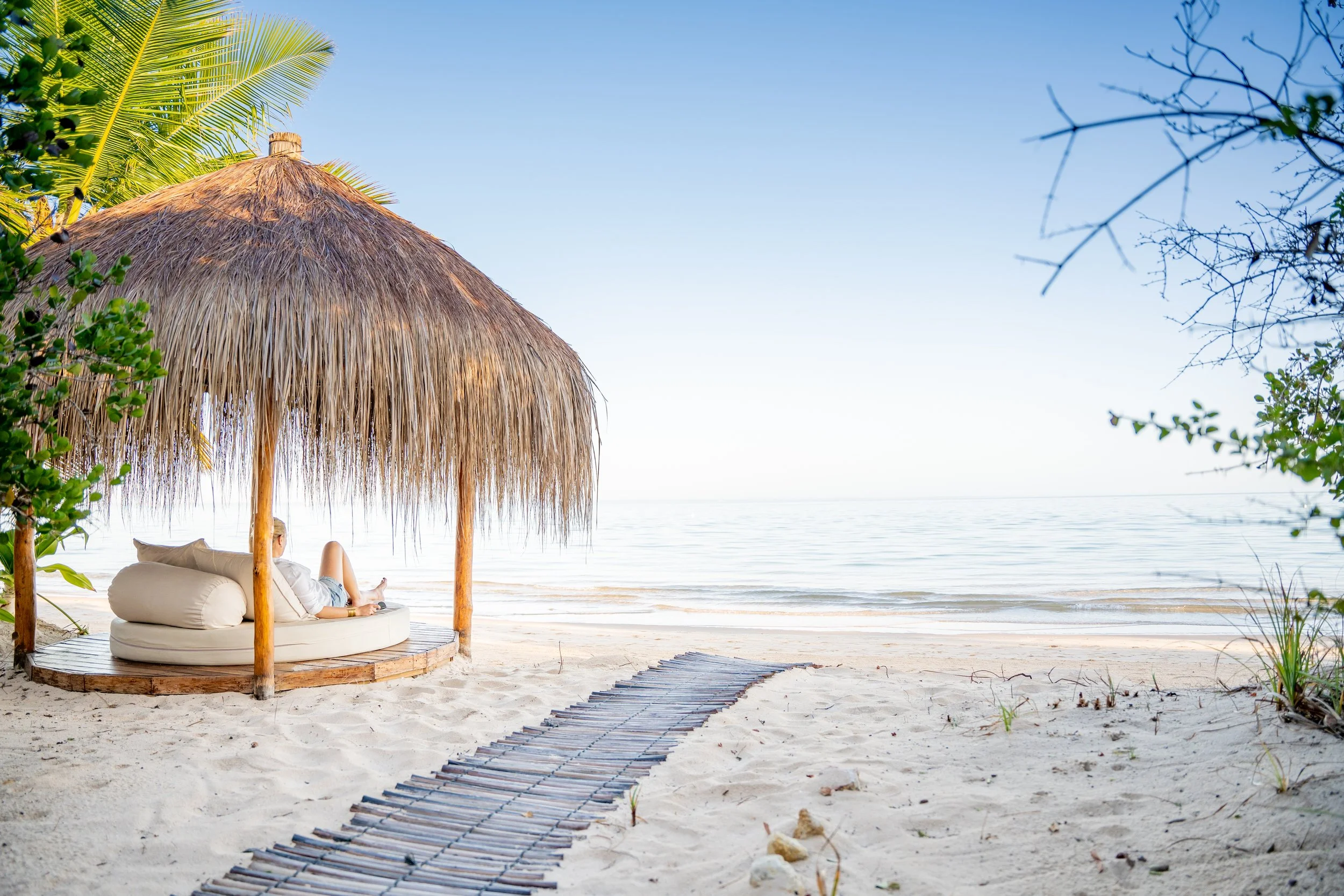 A person relaxing on a round cushioned seat under a thatched tiki hut on a sandy beach, with the ocean and a clear blue sky in the background.