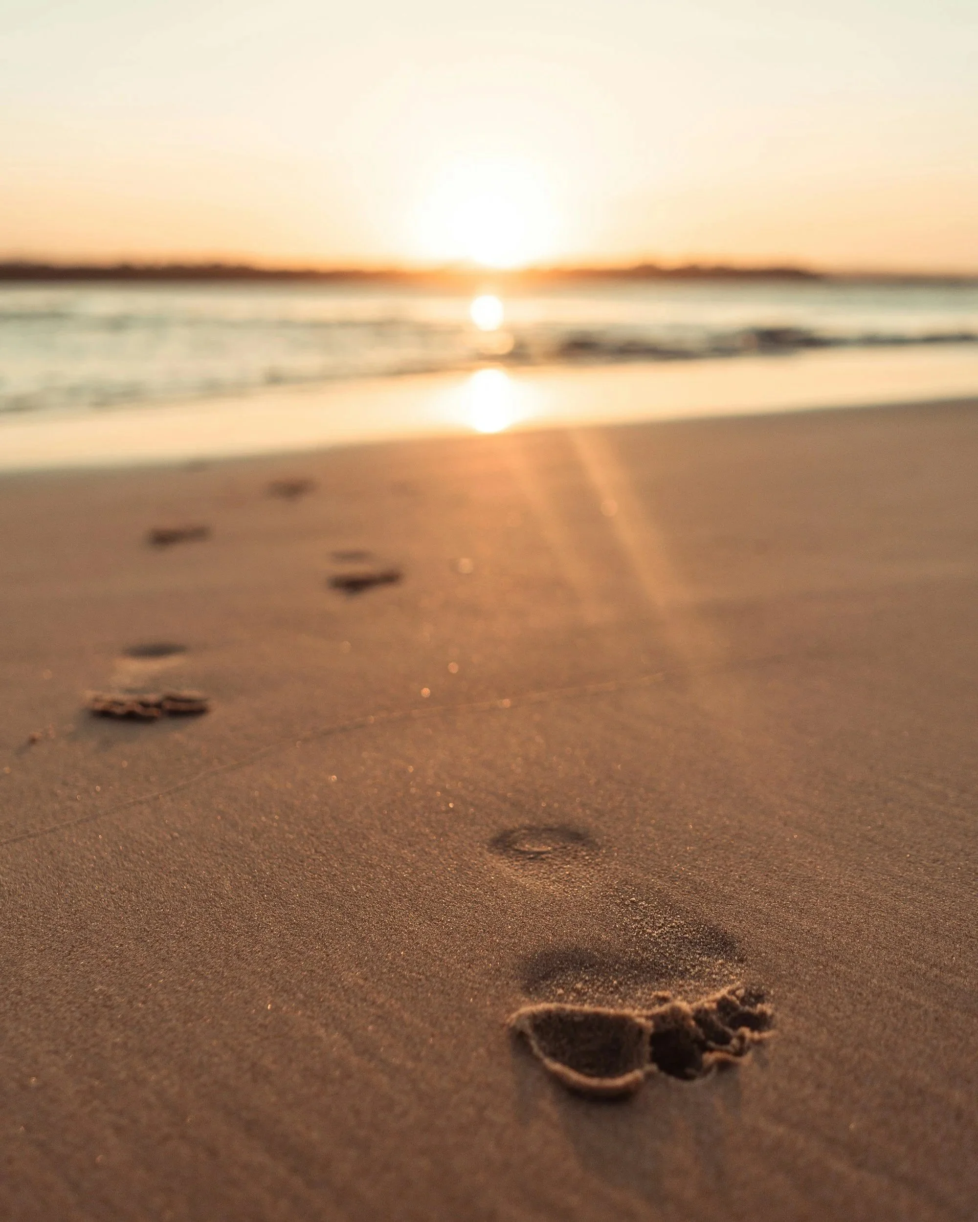 Footprints in wet sand leading towards the horizon at sunset, with the sun low in the sky over the ocean.