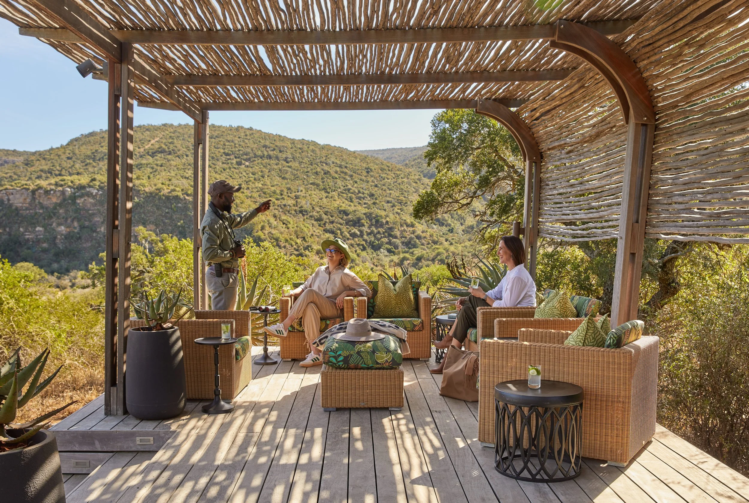 People sitting and talking on a covered outdoor wooden deck with mountain landscape in the background, using wicker furniture, with potted plants and drinks nearby.