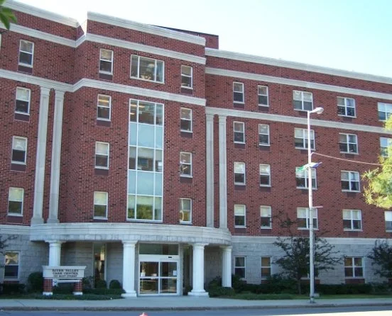 A multi-story brick building with large windows and a curved entrance supported by white columns, surrounded by trees and street lamps.
