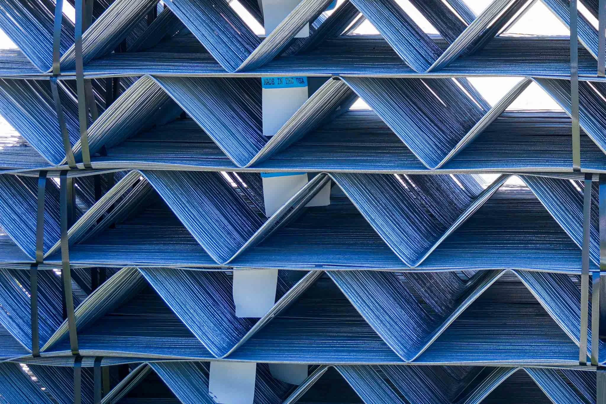 Stacked metallic blue pipes arranged horizontally in a metal storage rack, with a sign that reads 'Made in the USA' attached to the rack.