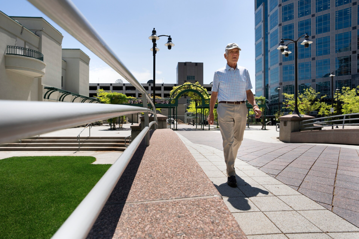 An elderly man walking on an urban sidewalk with modern high-rise buildings and green trees in the background, on a sunny day.