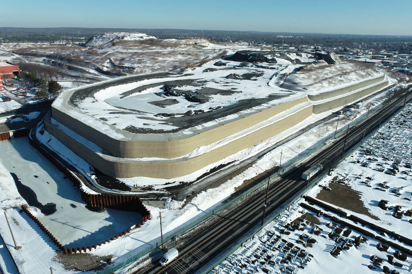 Aerial view of a large stadium with a snowy landscape, parking lot filled with snow-covered vehicles, and train tracks nearby during winter.