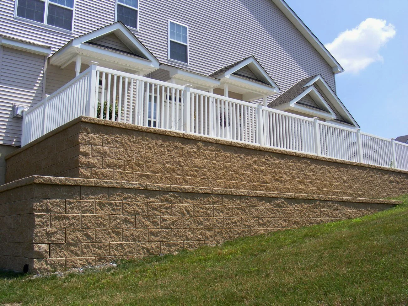 A house with a multi-level brick retaining wall, white railing, and siding, under a blue sky with a few clouds.
