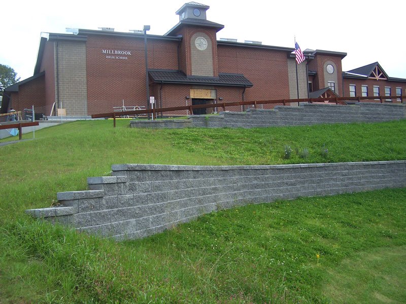 Front view of Millbrook High School building with brick exterior, American flag, and green grassy hill with concrete steps leading up to the entrance.