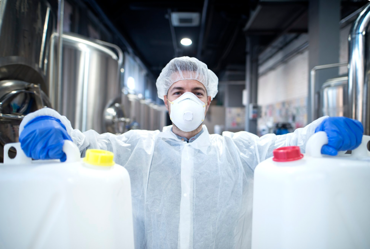 A worker in protective gear, including a face mask, hair cover, gloves, and a gown, standing between two large white containers with colorful caps, in an industrial or laboratory setting.