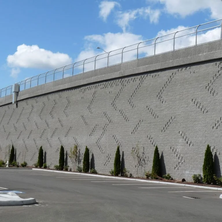 Concrete wall with a textured pattern, topped with a metal safety barrier, beside a parking lot with small trees and shrubs, under a partly cloudy sky.