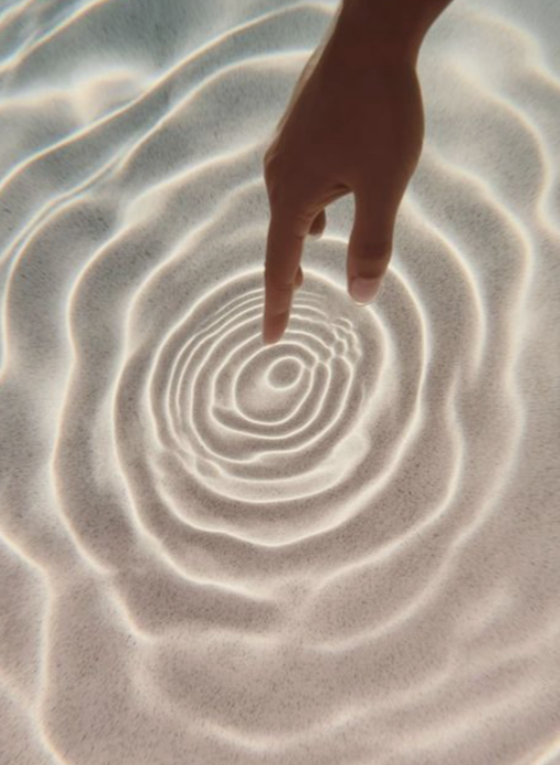 A hand with a finger pointing down, creating ripples in the sand that resemble concentric circles.