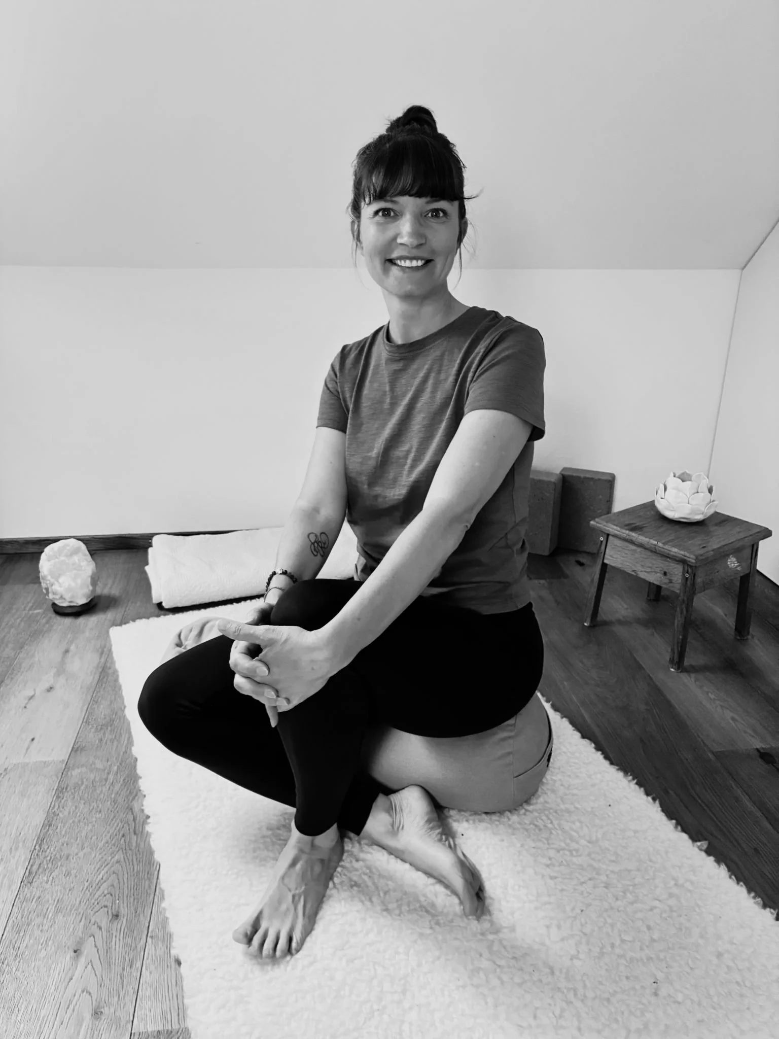 A woman with a bun hairstyle smiling, sitting on a cushion with her arms crossed, on a white plush rug. There are two rocks and a rolled towel on the rug, and a small side table with a white paper flower decoration in a room with light-colored walls and wood flooring.