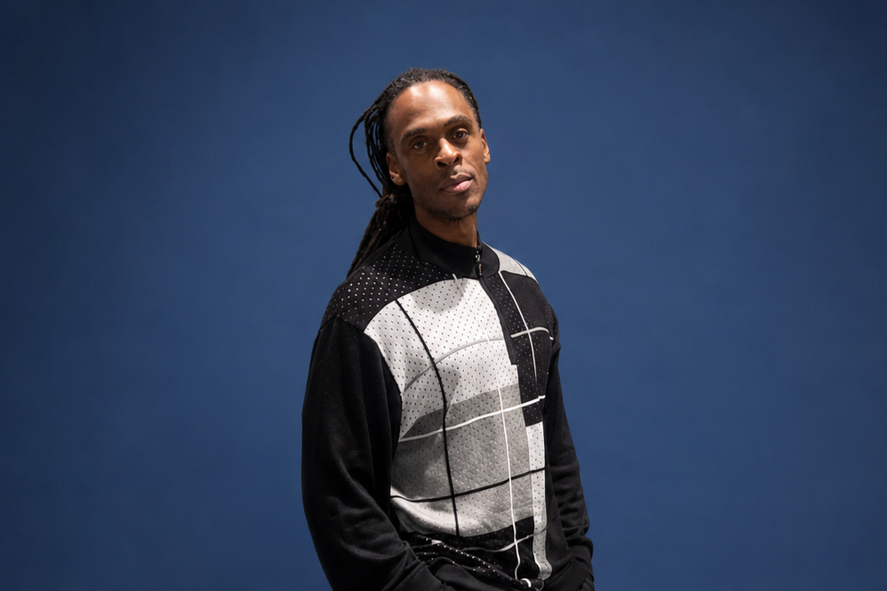 A man with long dreadlocks wearing a black and white patterned shirt stands against a blue background.