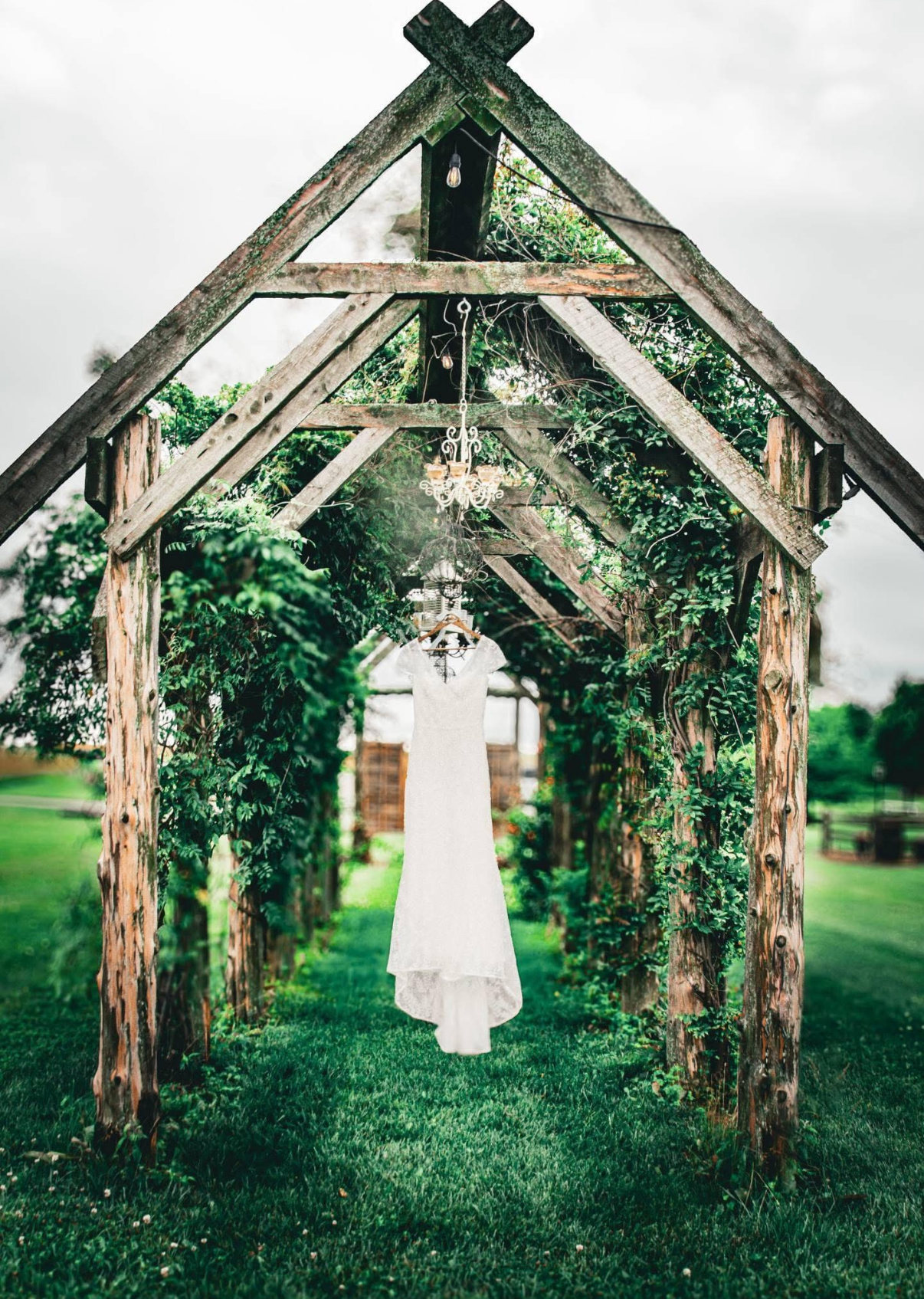 Outdoor wedding arch made of wooden beams with greenery and vines, featuring a hanging white wedding dress.