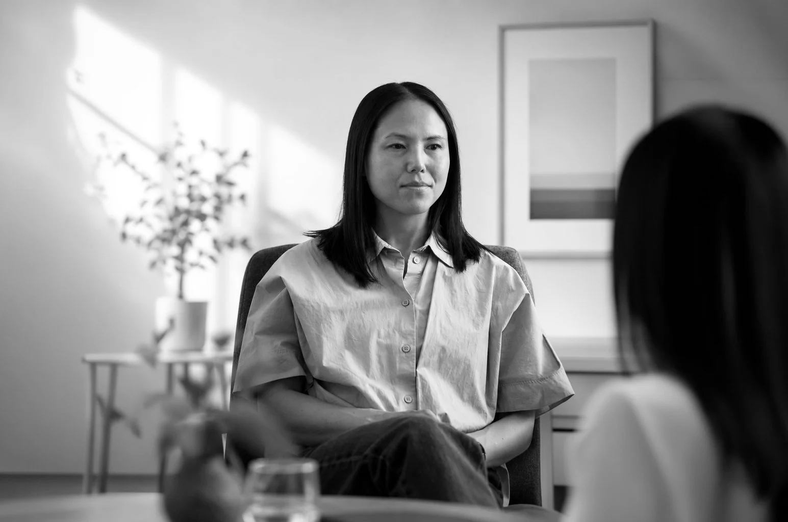 Sort-hvidt foto af Psykolog Maria Knudsen i samtale, en sidder foran, den anden er synlig fra ryggen, i et rum med et maleri på væggen og en plante i baggrunden.