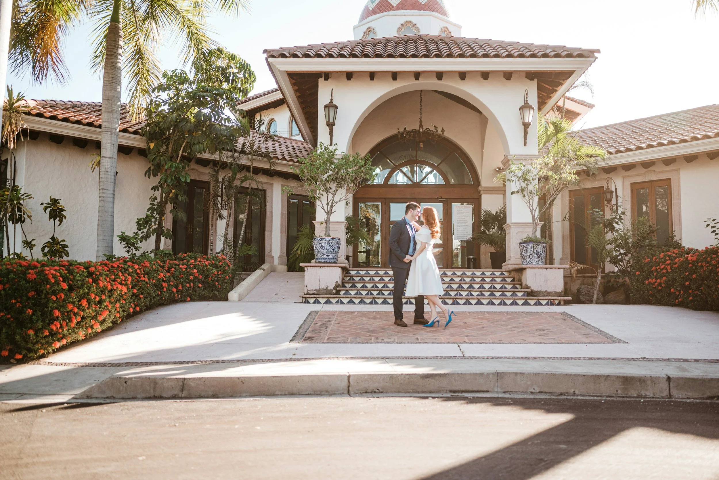 A wedding couple stands on steps in front of a Mediterranean-style building with a tiled roof, surrounded by palm trees and flowering bushes, during daytime.