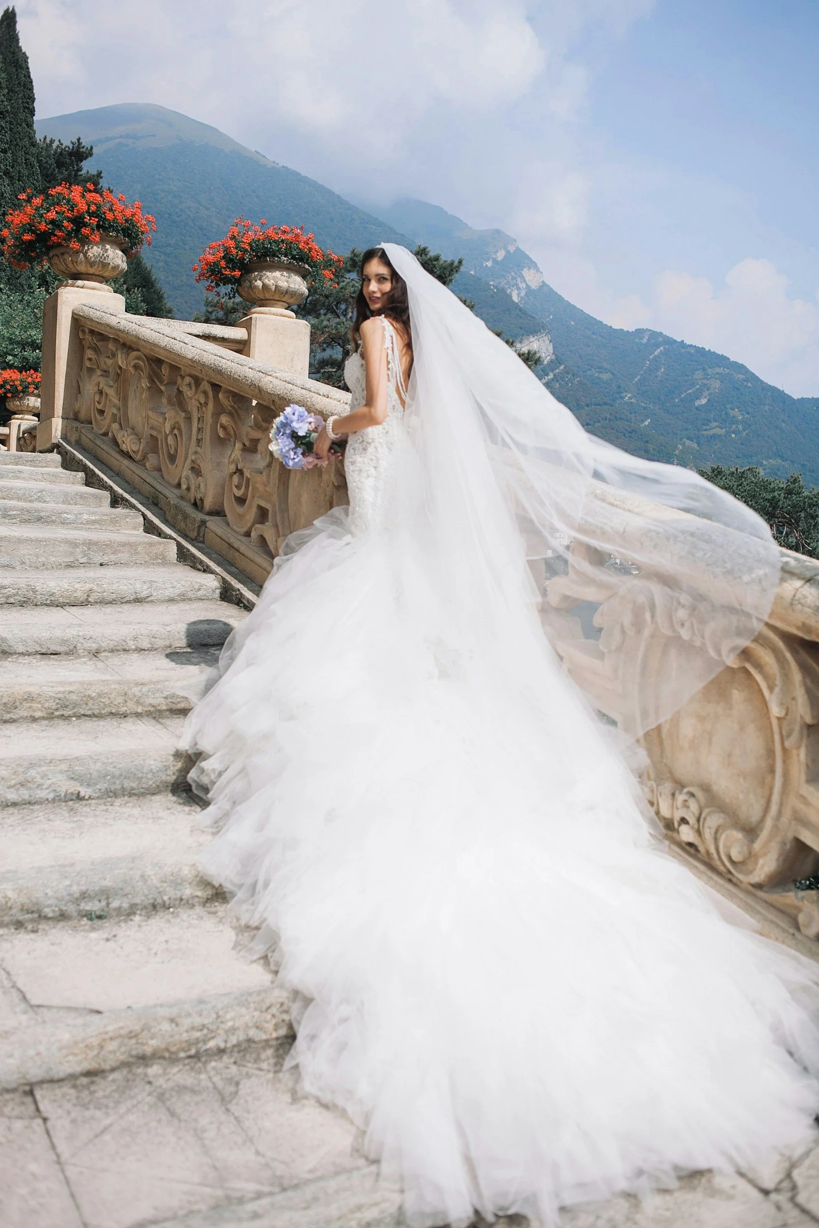 A bride in a white wedding gown with a long train standing on stone steps outdoors, holding a bouquet of purple flowers, with a mountain range and potted flowers in the background.