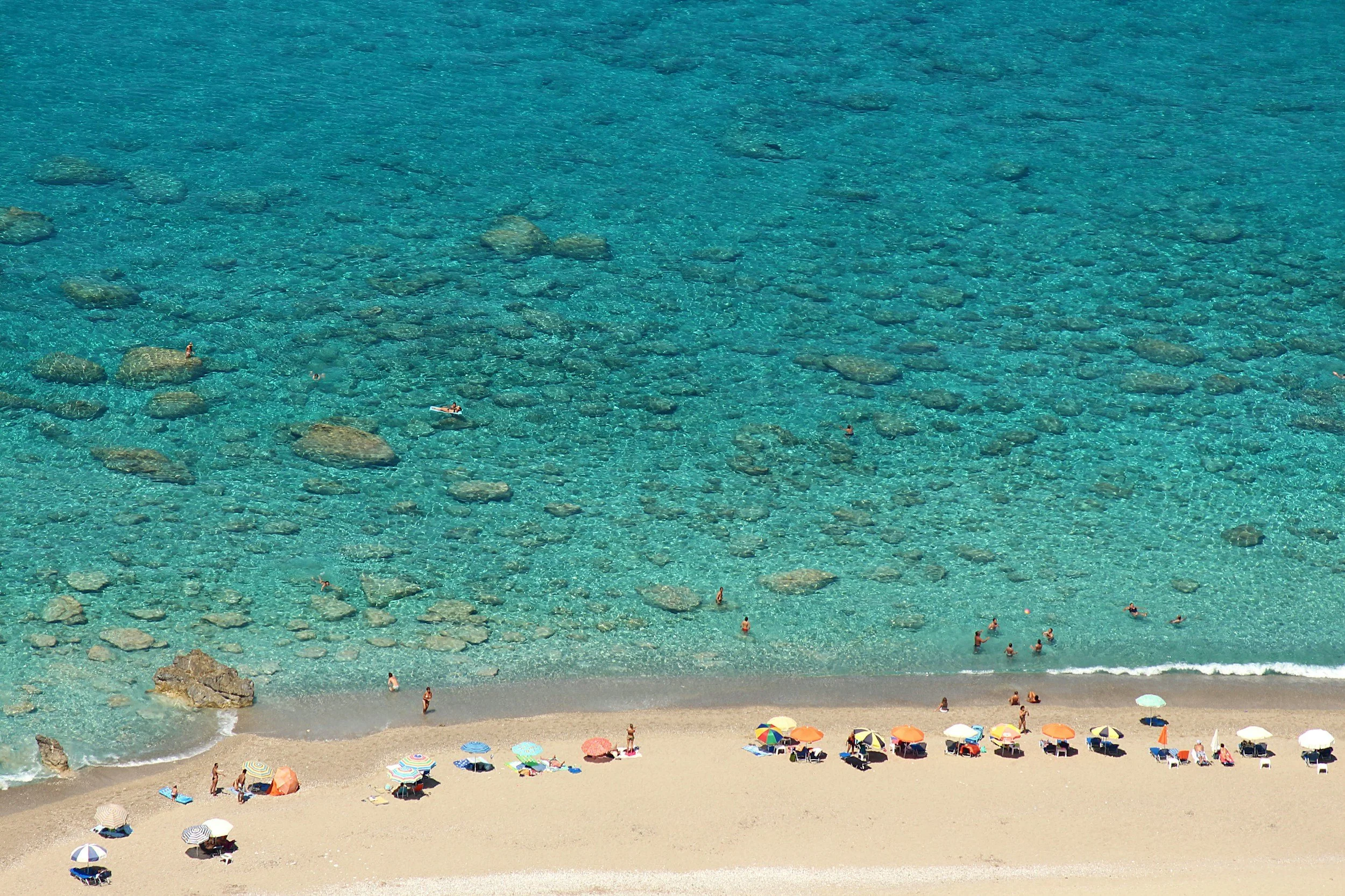 A beach scene with clear blue water, some large rocks in the water, and a sandy shore with colorful umbrellas and people enjoying the beach.