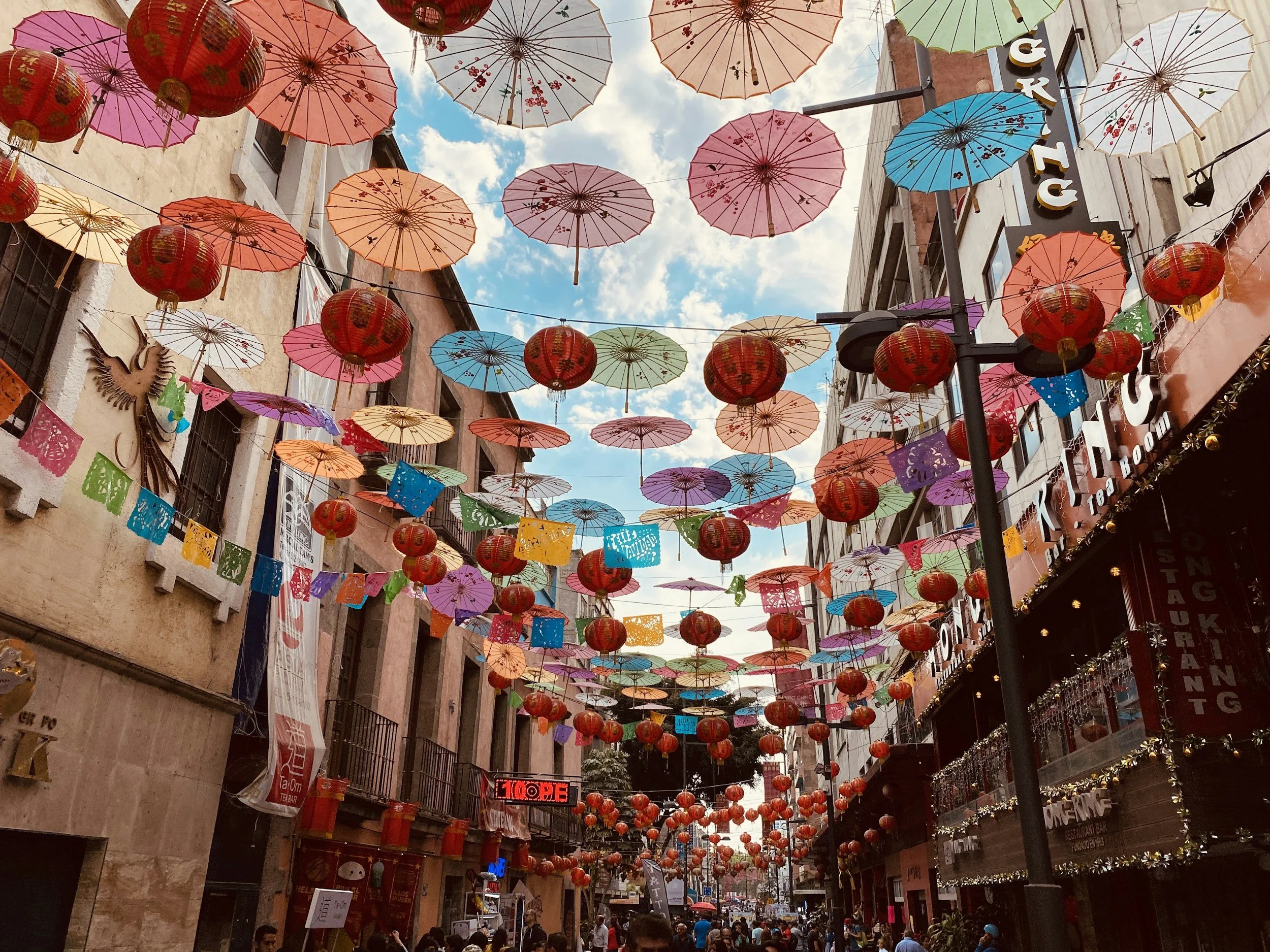 Colorful umbrellas and lanterns hanging above a busy street during daytime, with buildings on either side and a partly cloudy sky.