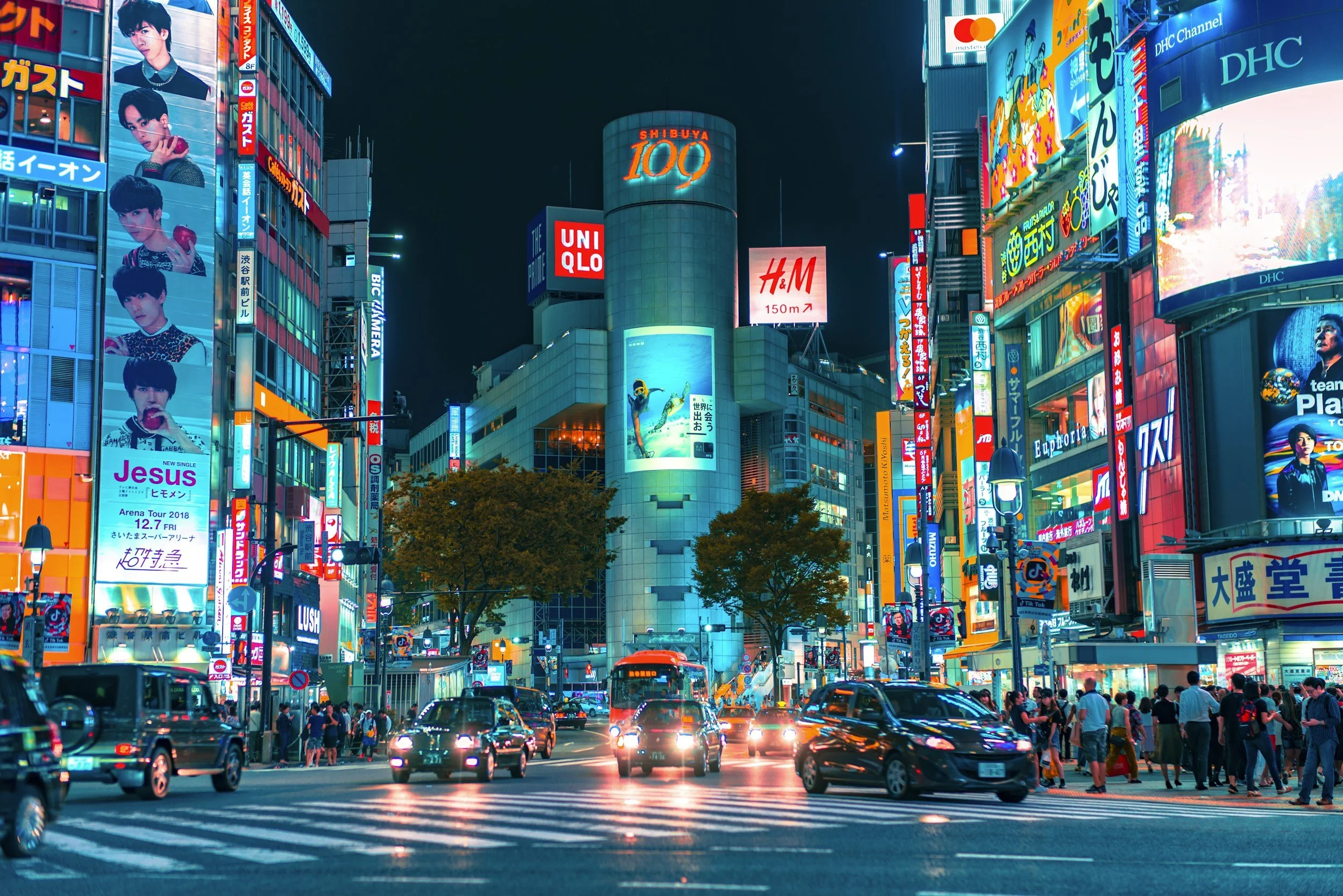 Nighttime view of Shibuya Crossing in Tokyo, Japan, with crowds of people crossing the street, surrounded by brightly lit billboards and advertisements on tall buildings.