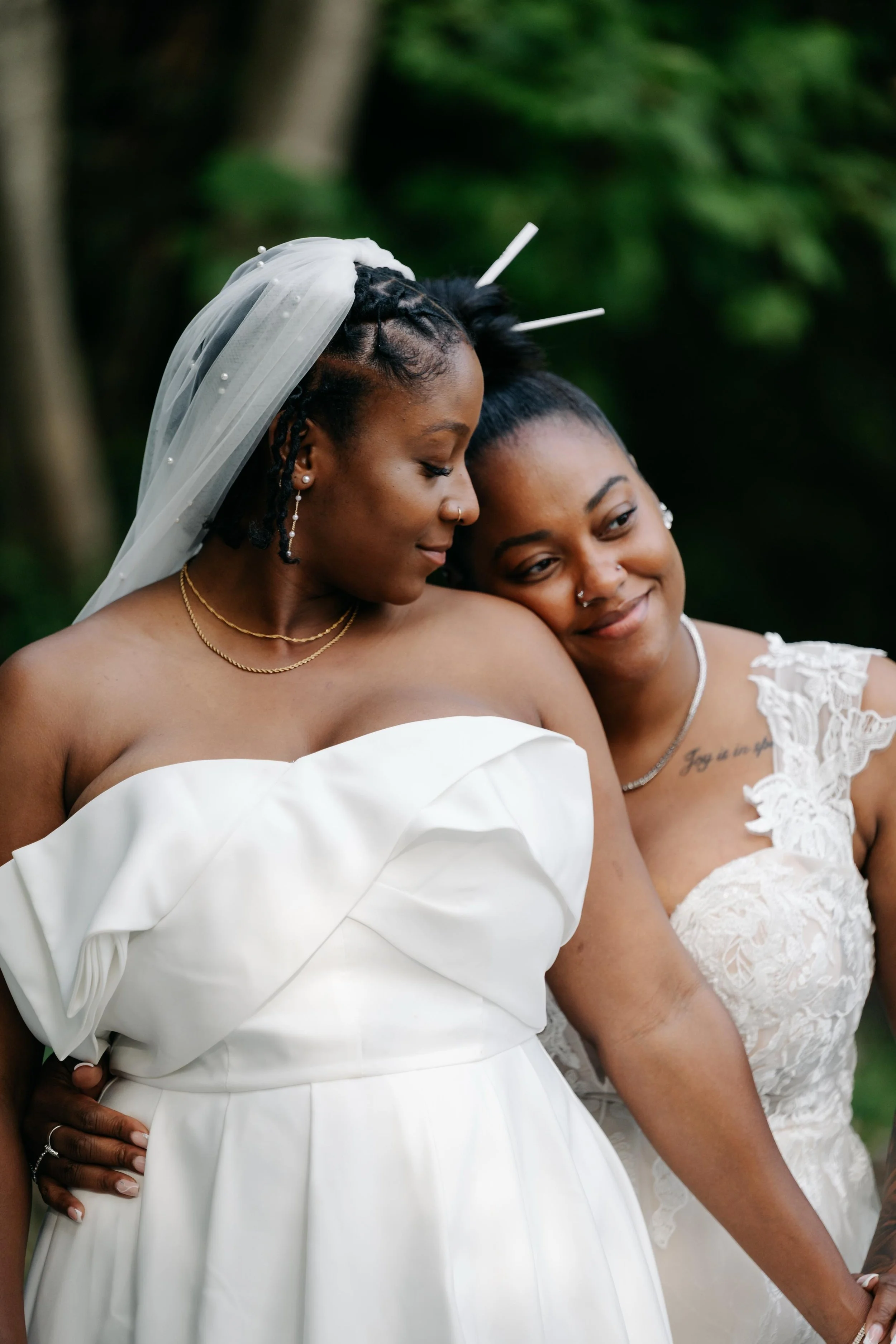 Two Black women embracing in white wedding attire, with one wearing a beaded veil, outdoors.