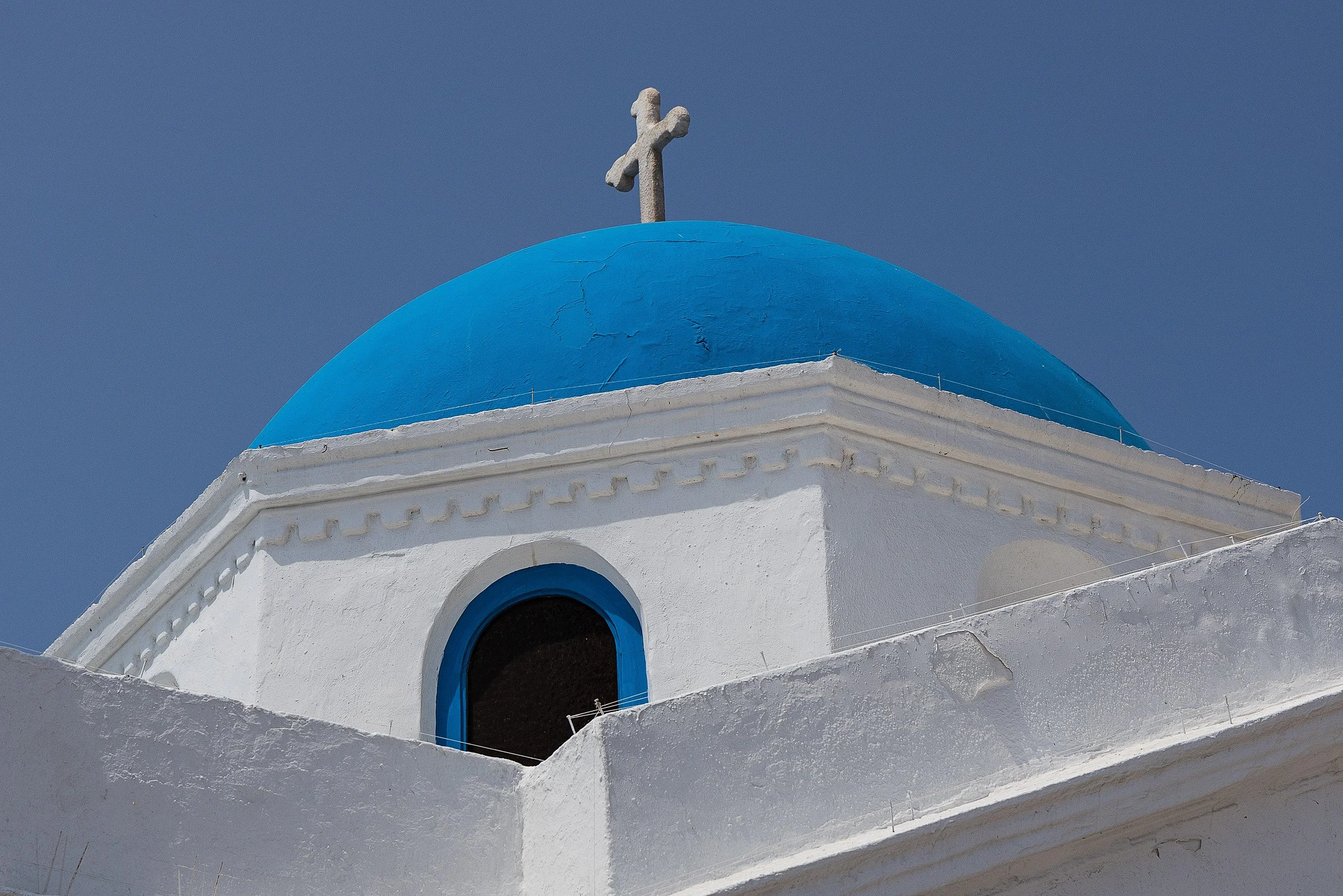 A white building with blue accents and a blue dome roof topped with a cross, typical of Greek Orthodox churches, against a clear blue sky.
