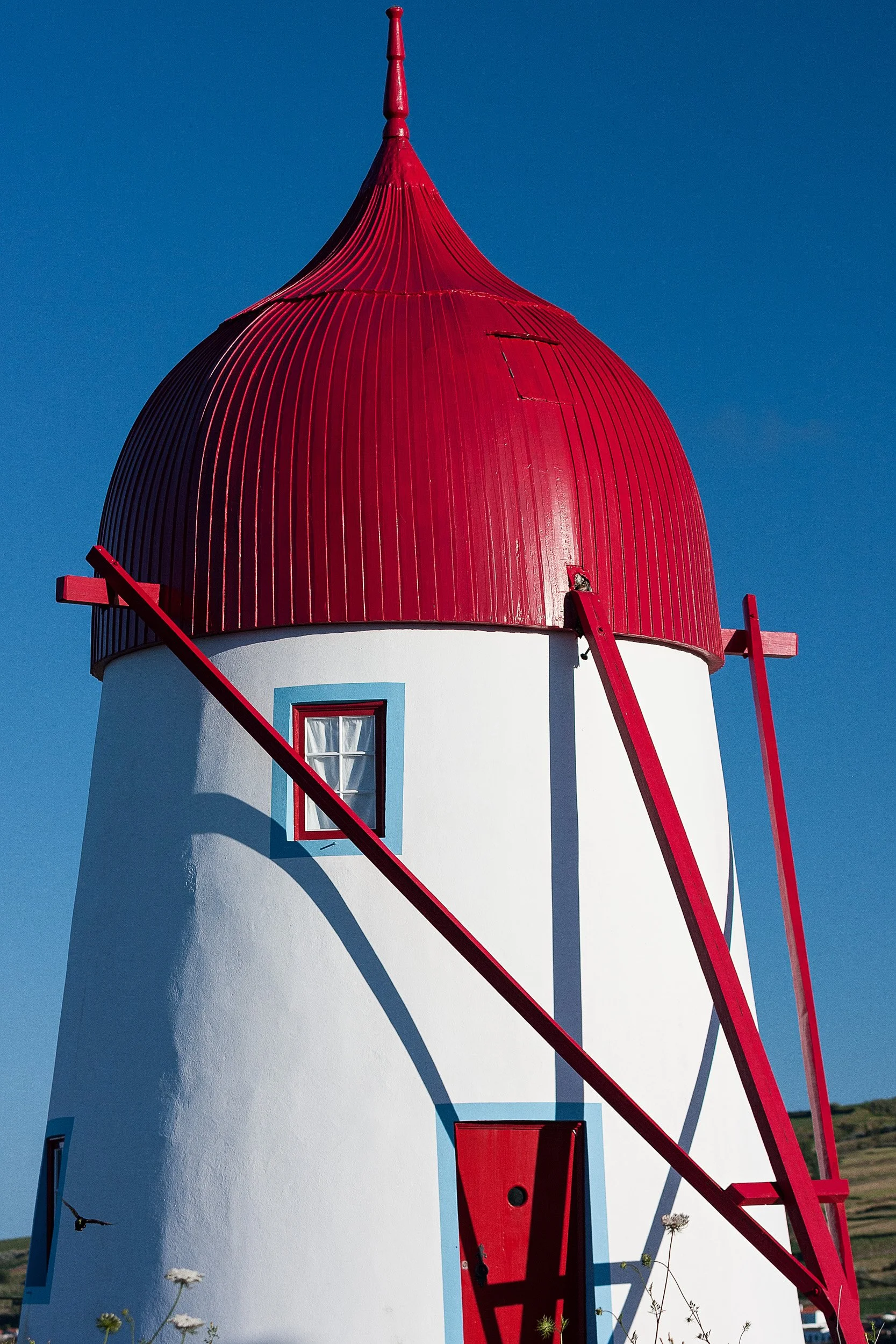 A white lighthouse with a red, conical cap and red details, including a door and supporting beams, against a clear blue sky.