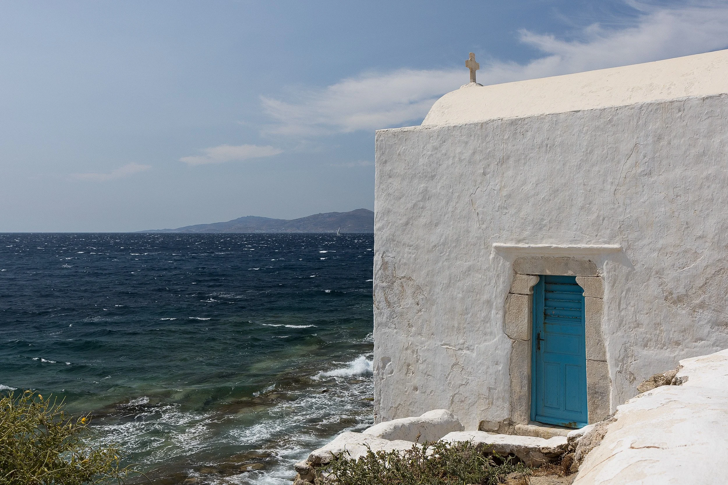 A white stucco building with a blue shuttered window and a cross on top, located beside the ocean with waves and a distant island or landmass in the background under a partly cloudy sky.