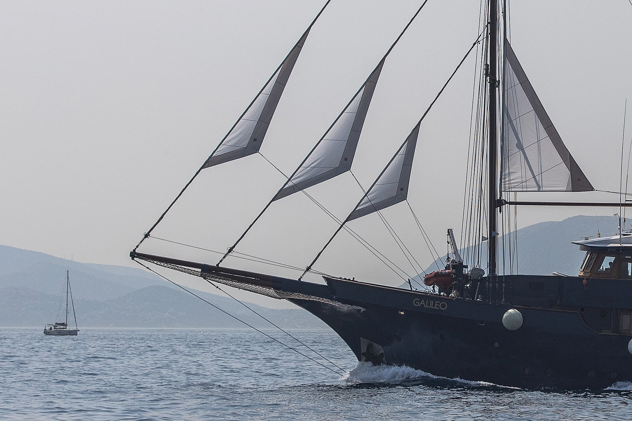 A large sailboat named Galileo with modern sails cutting through the water with another sailboat in the background and distant mountains under an overcast sky.