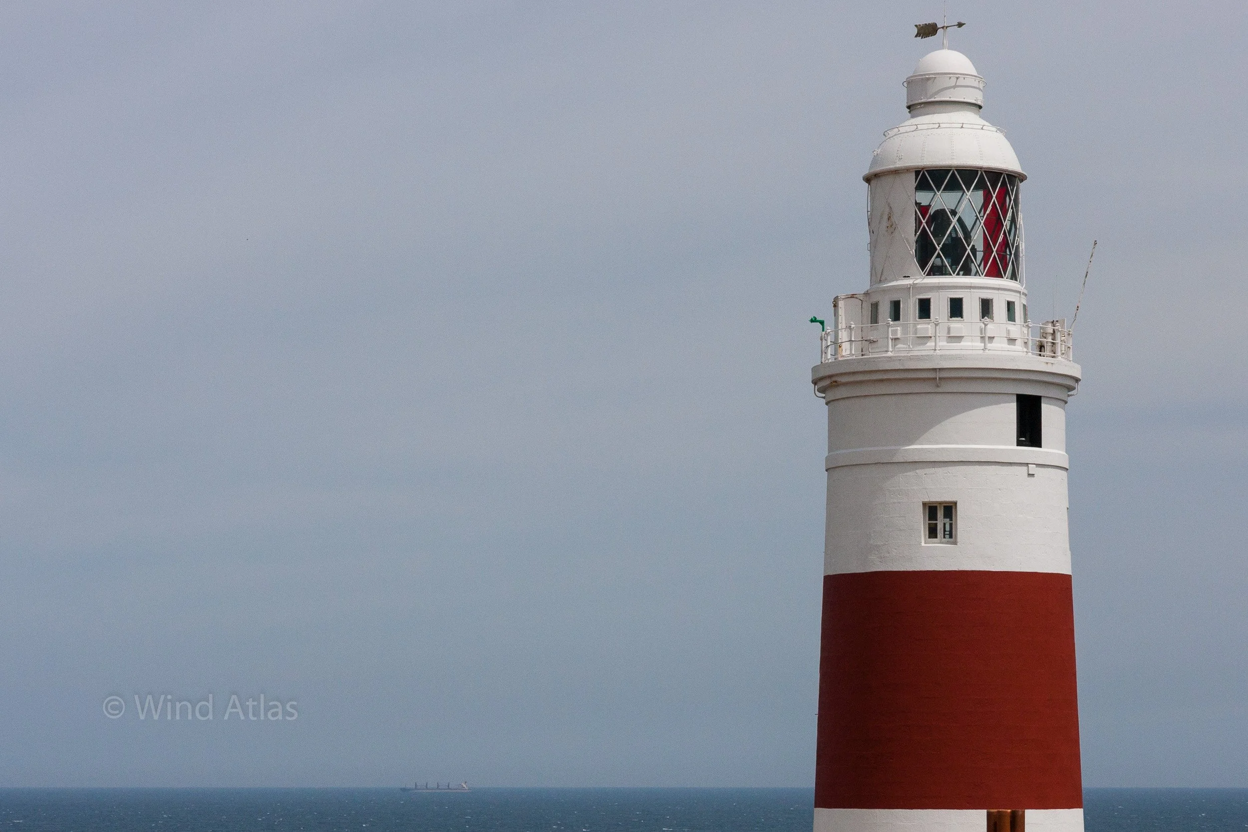 A white lighthouse with a red band near the bottom and black window panes, standing against a cloudy sky over the ocean with a ship visible in the distance.
