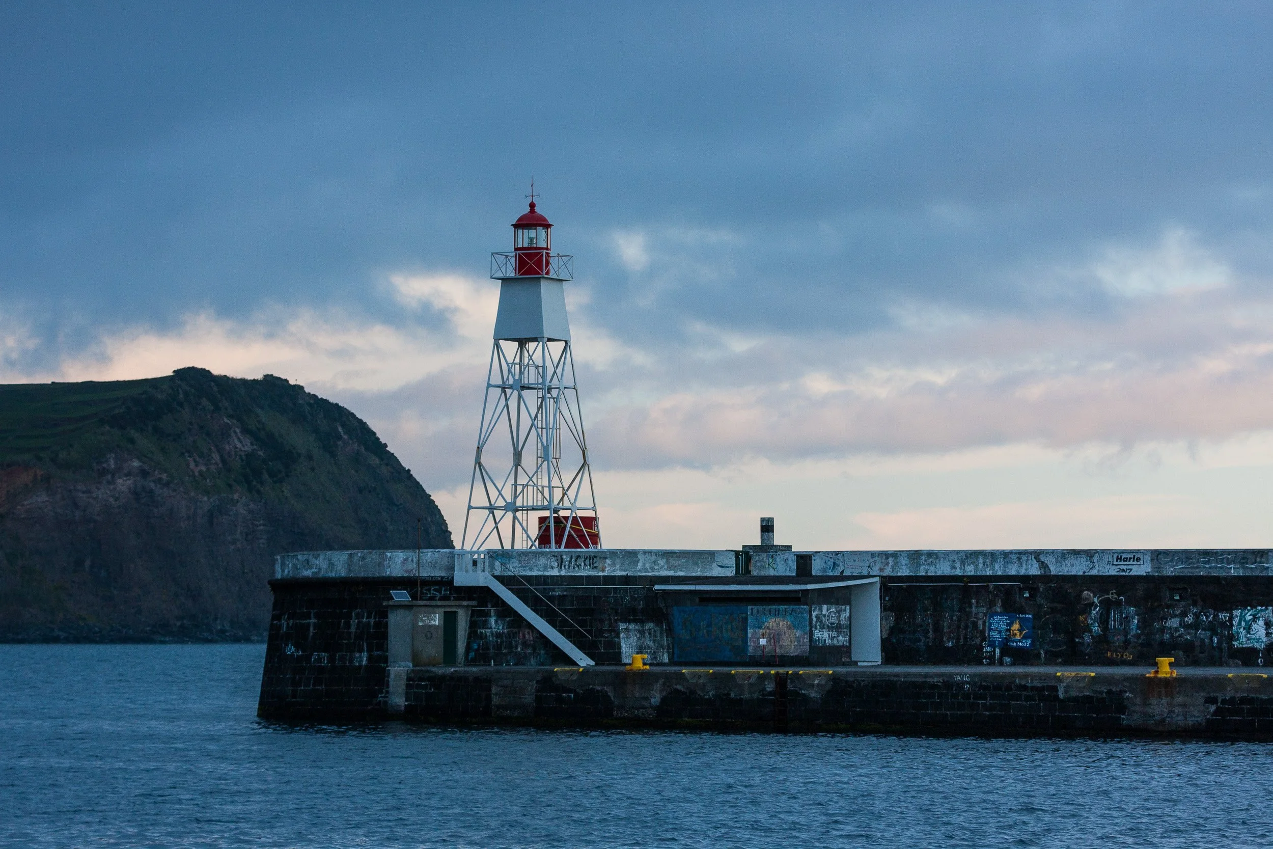 A lighthouse on a pier with graffiti, with a mountain in the background during cloudy weather.