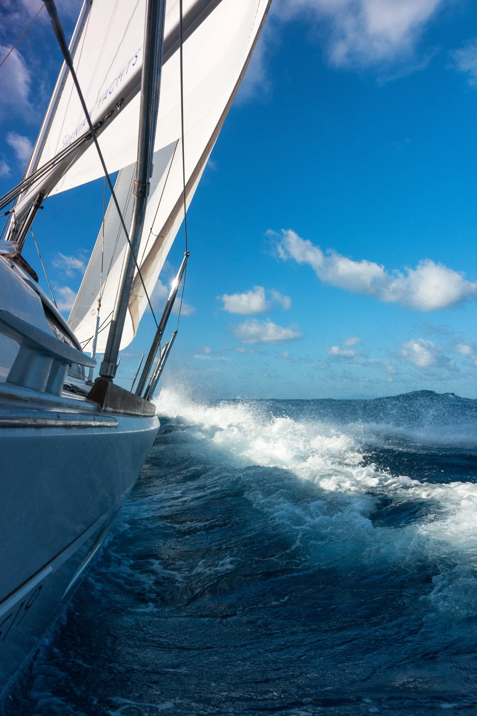 View from the side of a sailboat moving through the water on a sunny day, with waves splashing behind, blue sky, and a few clouds.