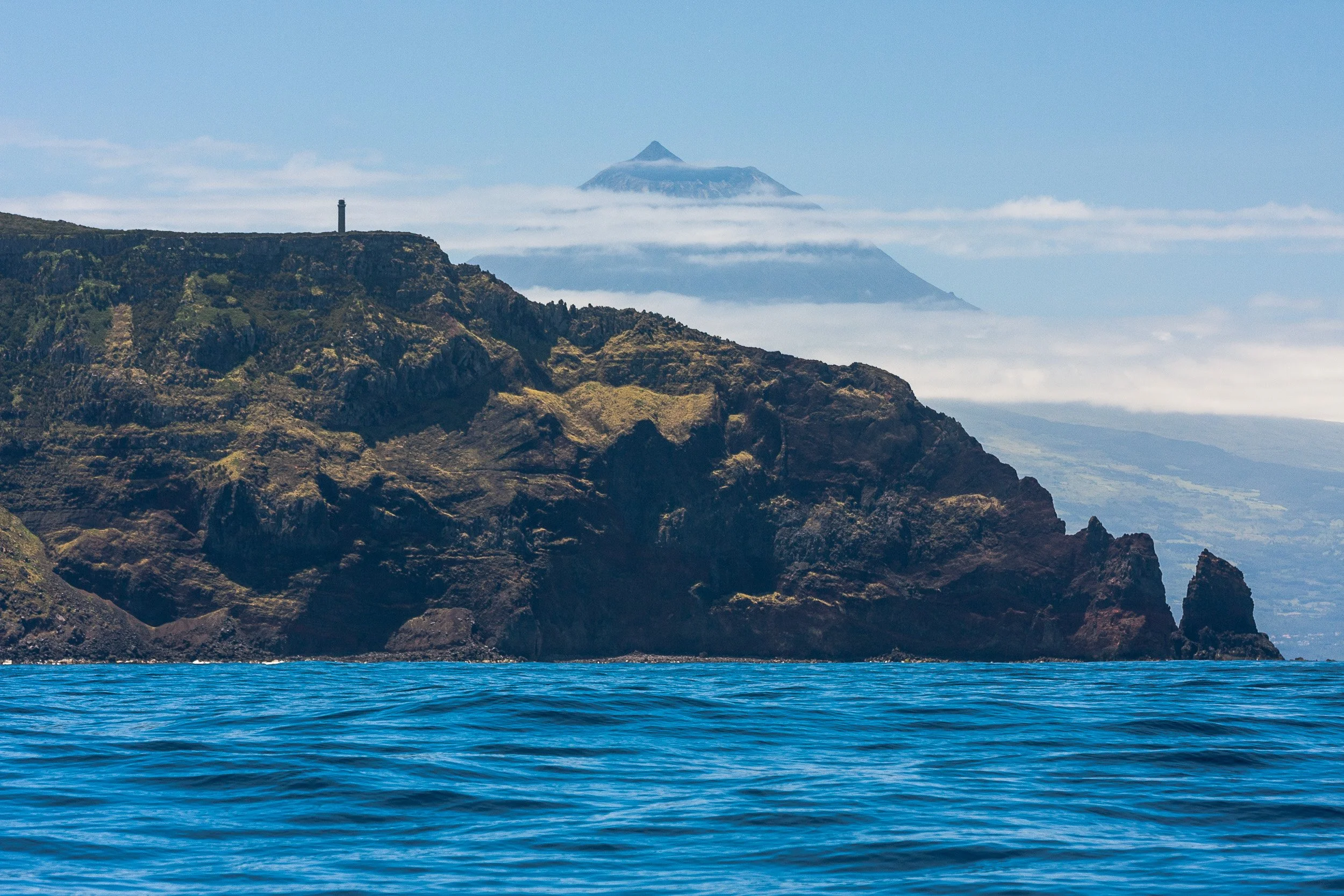 Ocean with blue water in the foreground, rocky coastline with cliffs in the middle ground, and a mountain with clouds surrounding it in the background.