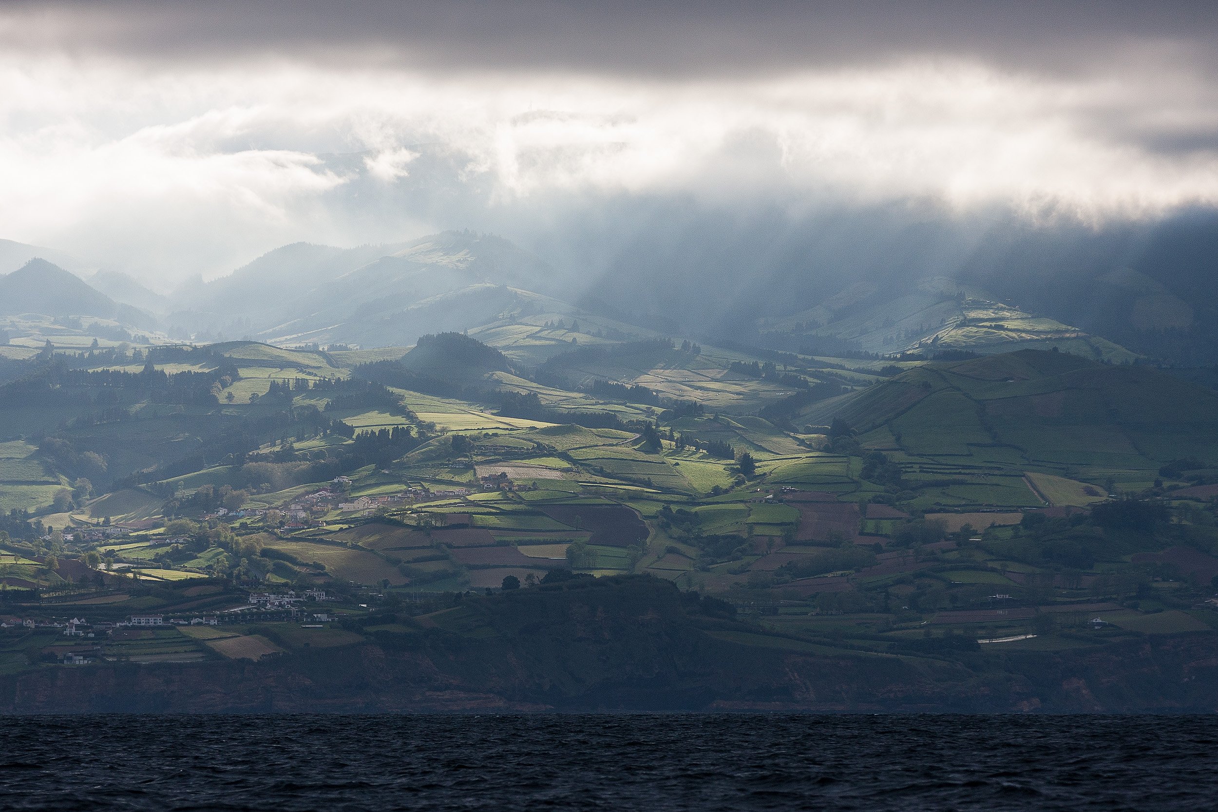 Scenic view of a landscape with rolling green hills, patchwork farmland, and mountains under a sky with clouds.