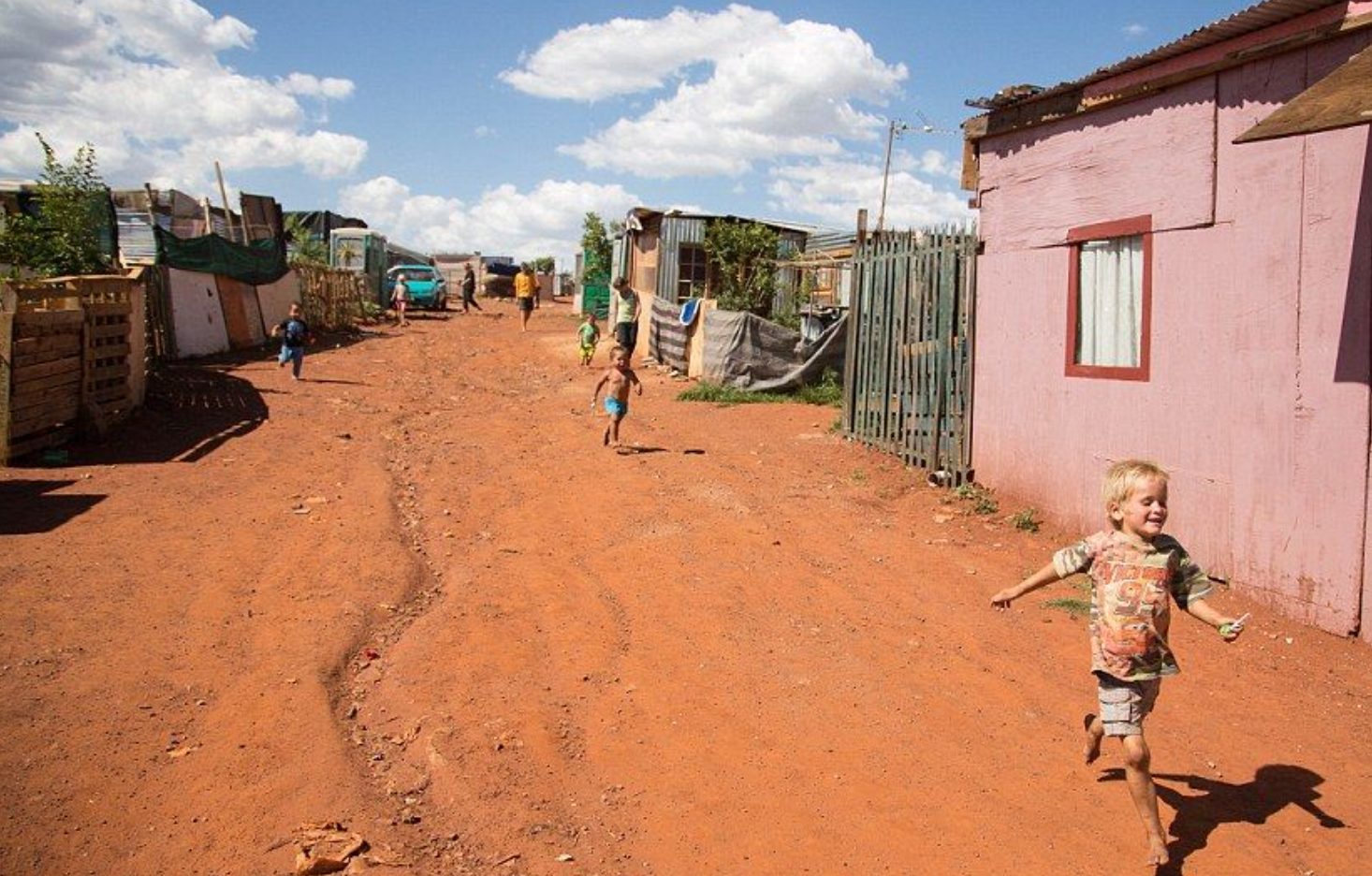 A dirt road in a low-income neighborhood with children playing, houses made of wood and metal, and a bright blue sky.