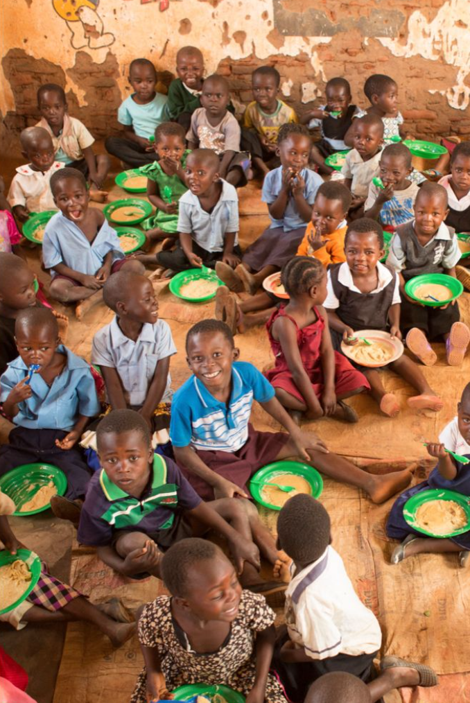 Group of African children sitting on a floor, eating from green plates, with a rustic wall in the background.