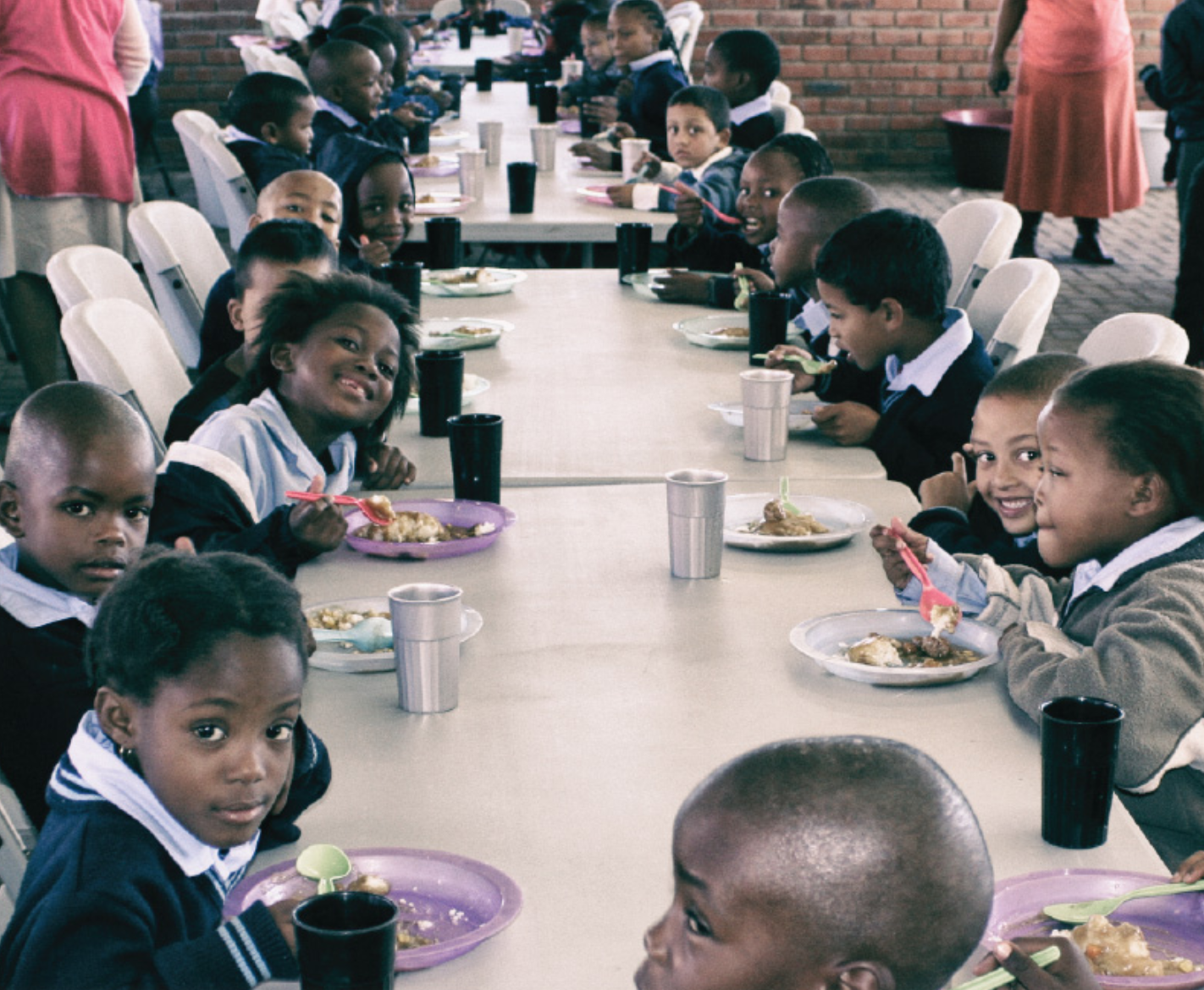 Children sitting at a long table in a cafeteria, eating from plates with plastic utensils, some smiling and looking at the camera.