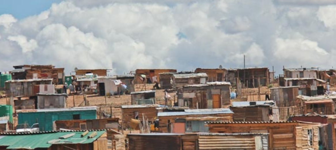 A wide view of a densely populated informal settlement with makeshift houses made of various materials, situated on a hillside under a partly cloudy sky.