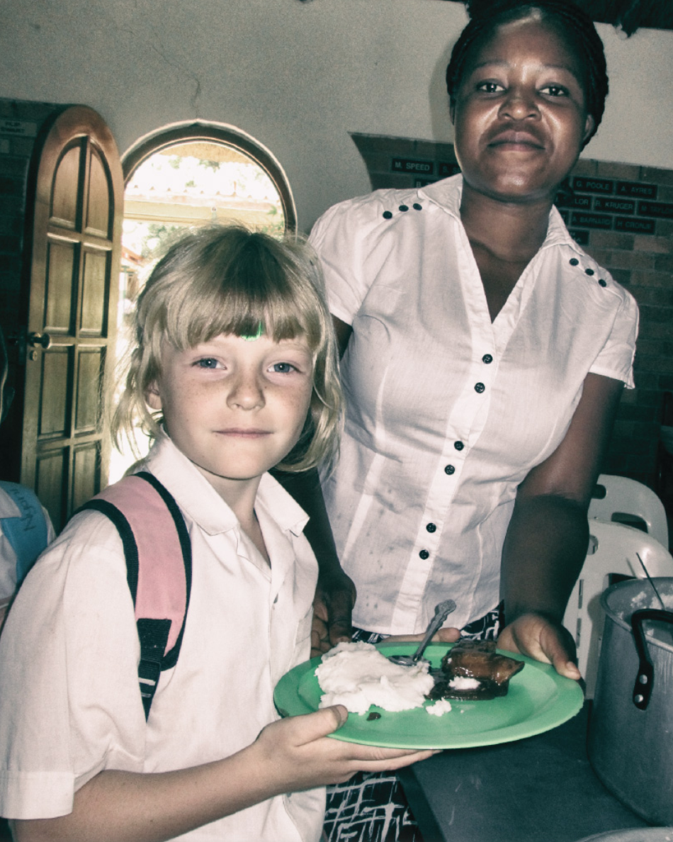 Young girl with blonde hair and a pink backpack holding a green plate with cake and ice cream, standing next to a woman with short braided hair in a white button-up shirt inside a building with a brick wall and an open window.