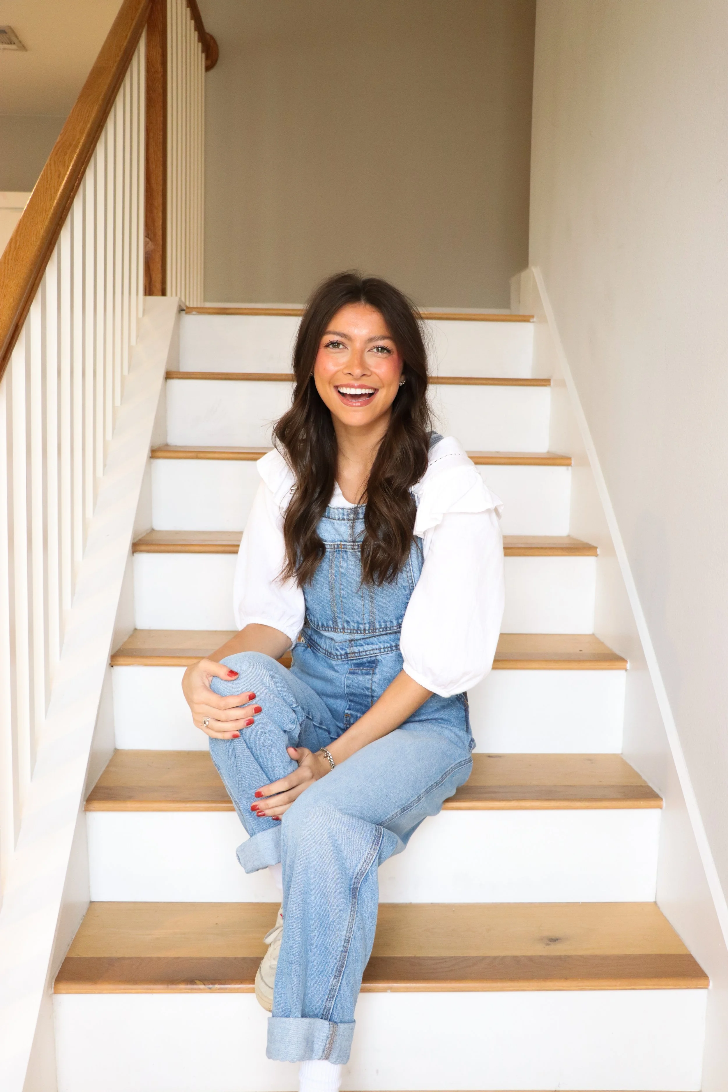 A woman with long brown hair sitting on wooden stairs, smiling and looking at the camera, wearing a white blouse and denim overalls.