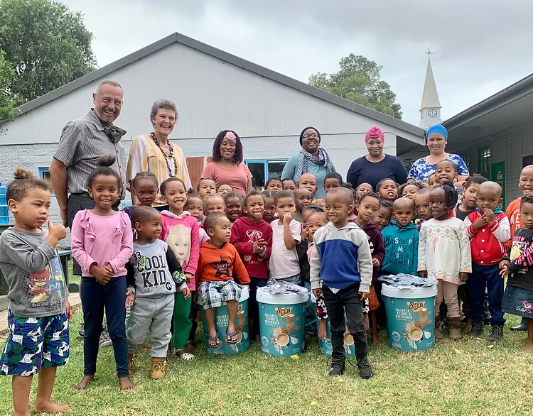 Group of children and adults outdoors in front of a building with a church steeple in the background. The children are smiling, some sitting on large blue buckets, and the adults are standing behind them.