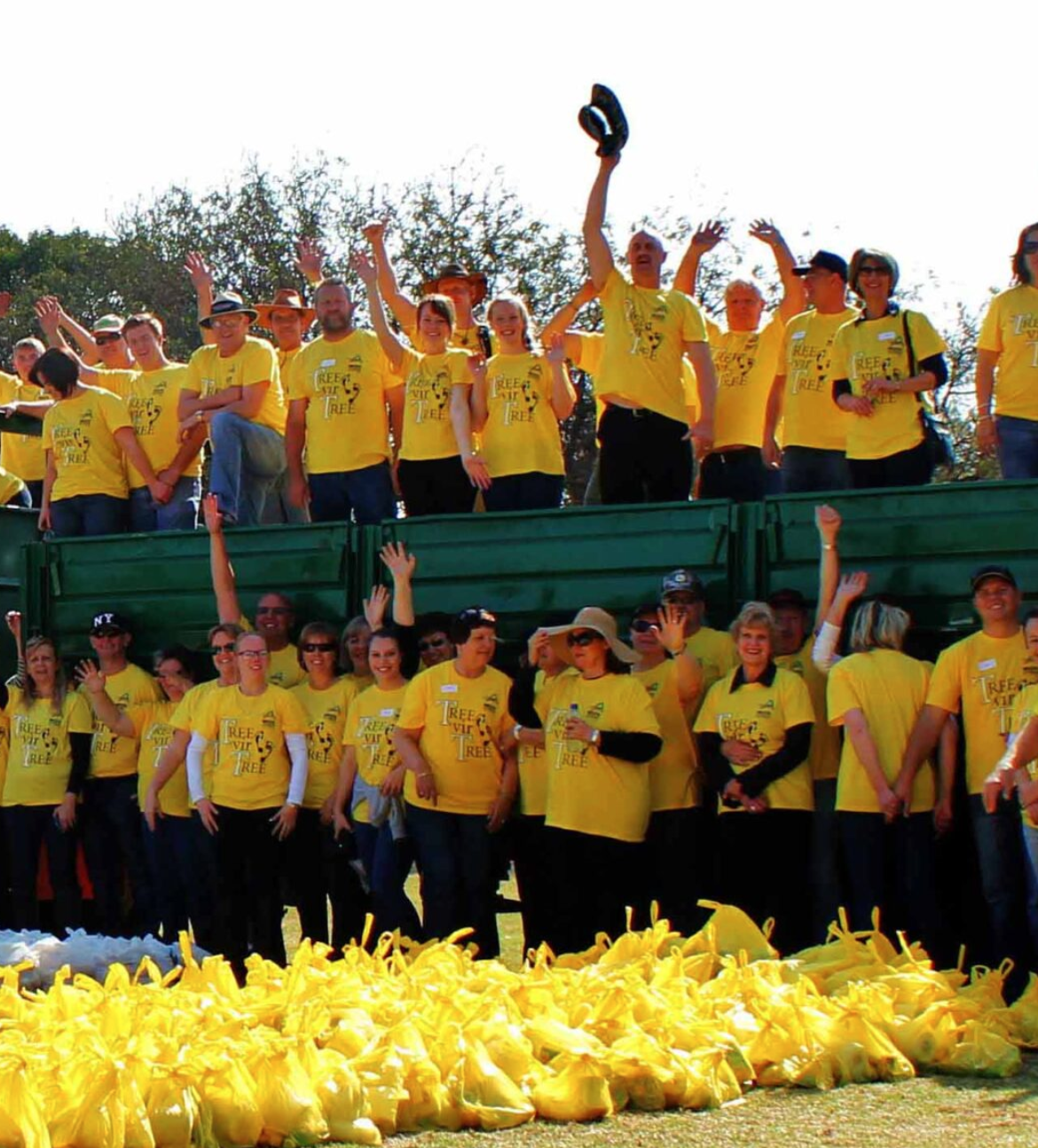 A large group of people wearing yellow T-shirts gathered outdoors, some standing on a green platform and others on the ground, with yellow bags of what appears to be flowers or fabric in the foreground. Many are smiling, waving, or raising their hands, participating in a cheerful event.