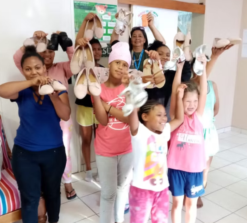 Group of children and an adult smiling and holding up shoes and slippers in an indoor setting