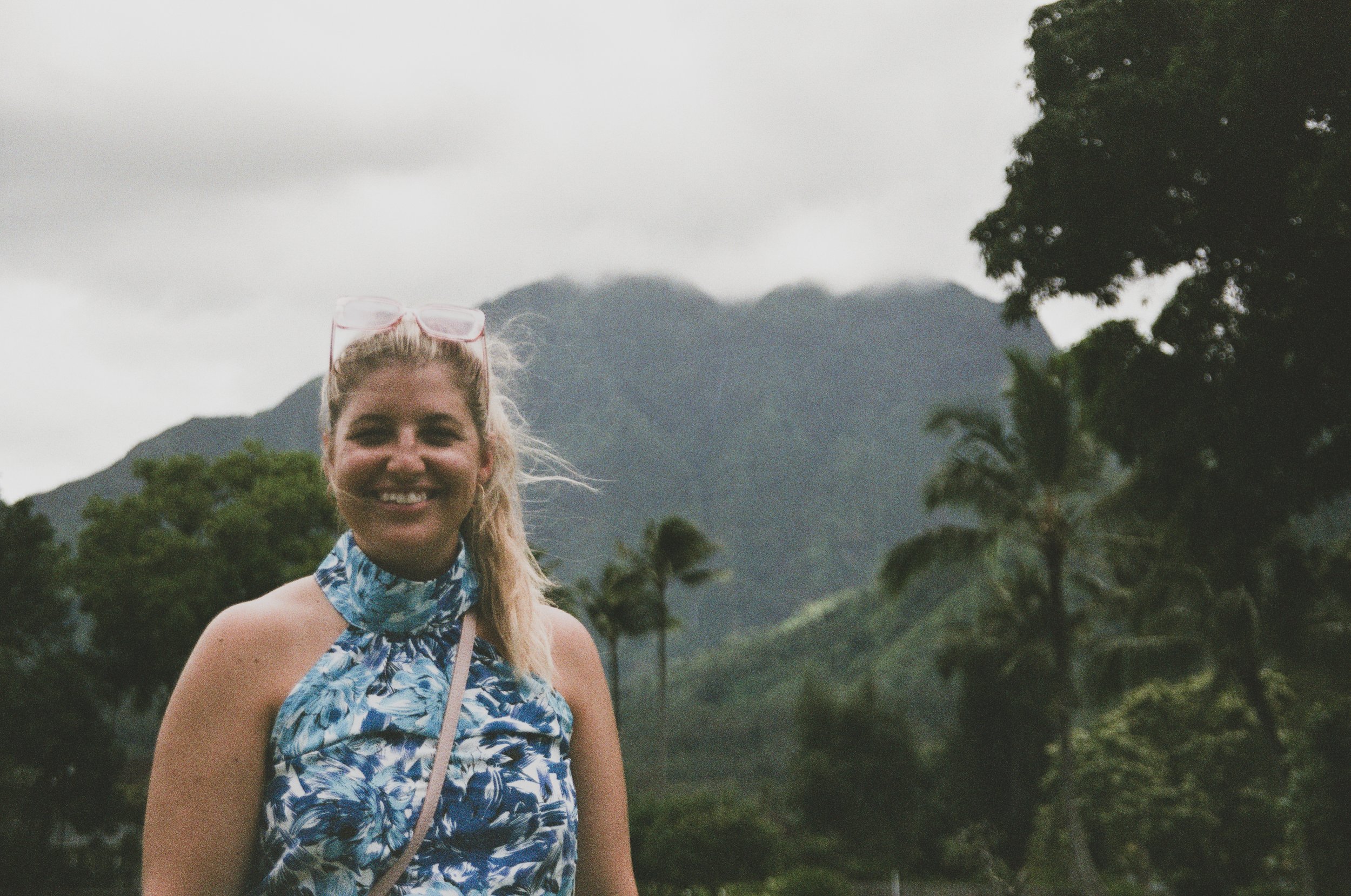 A woman smiling outdoors with mountains, trees, and cloudy sky in the background.
