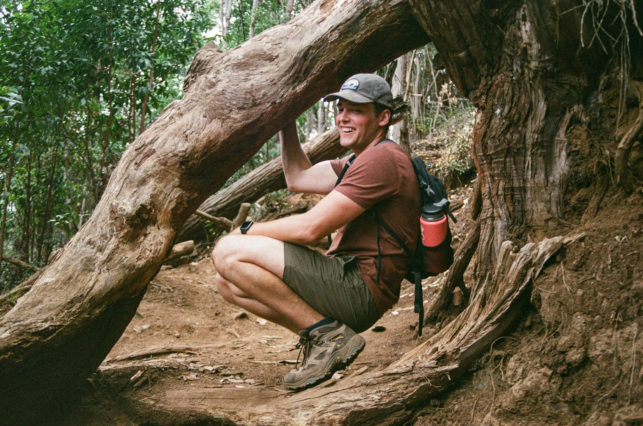 A man wearing a brown t-shirt, gray shorts, hiking shoes, and a baseball cap is smiling and crouching next to a fallen tree in a forest, with his hands on the tree.