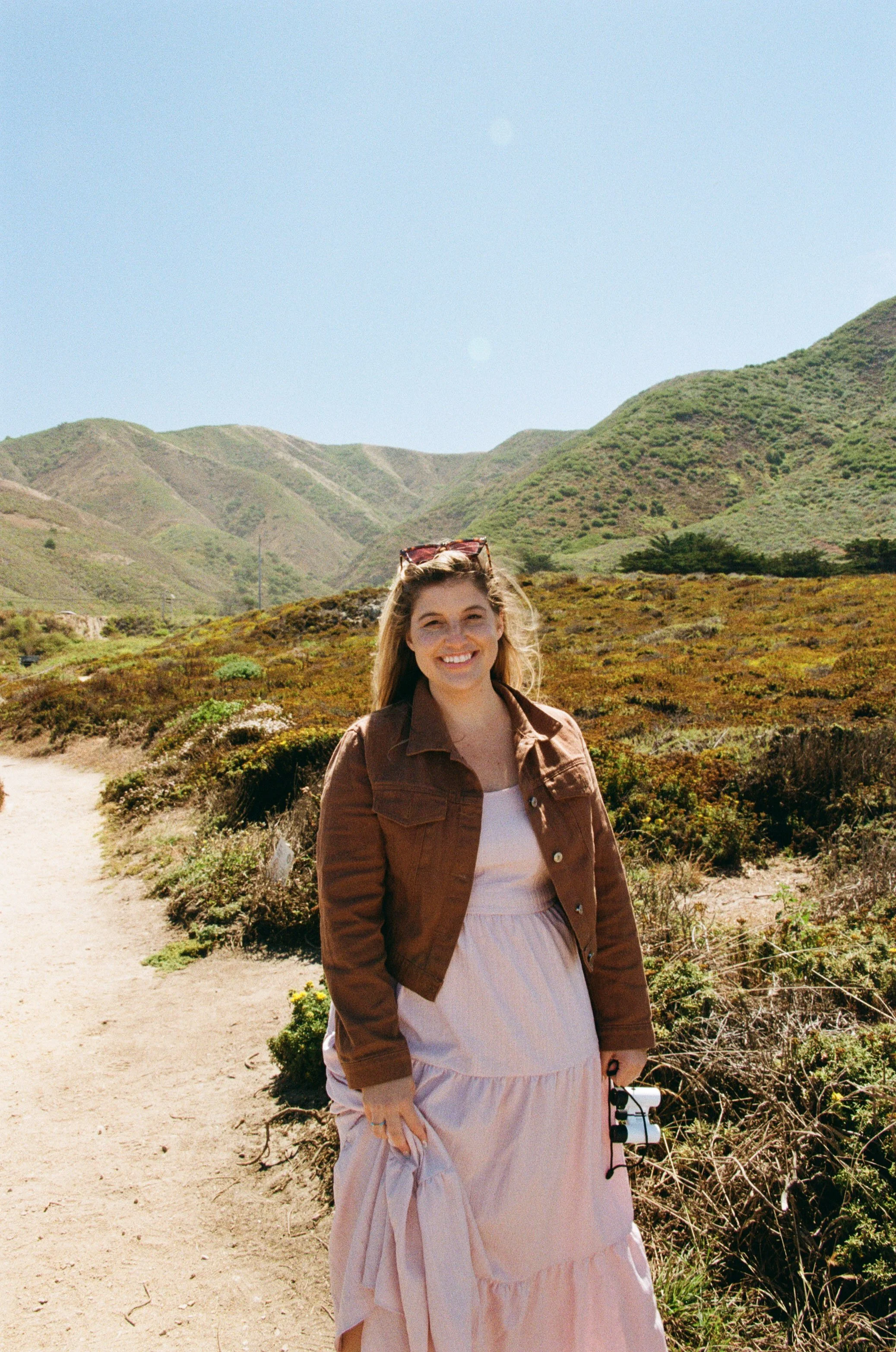 A young woman with blonde hair and sunglasses on her head, smiling and standing on a dirt path in a hilly, green landscape under a clear blue sky. She wears a brown jacket over a white top and a pink skirt.
