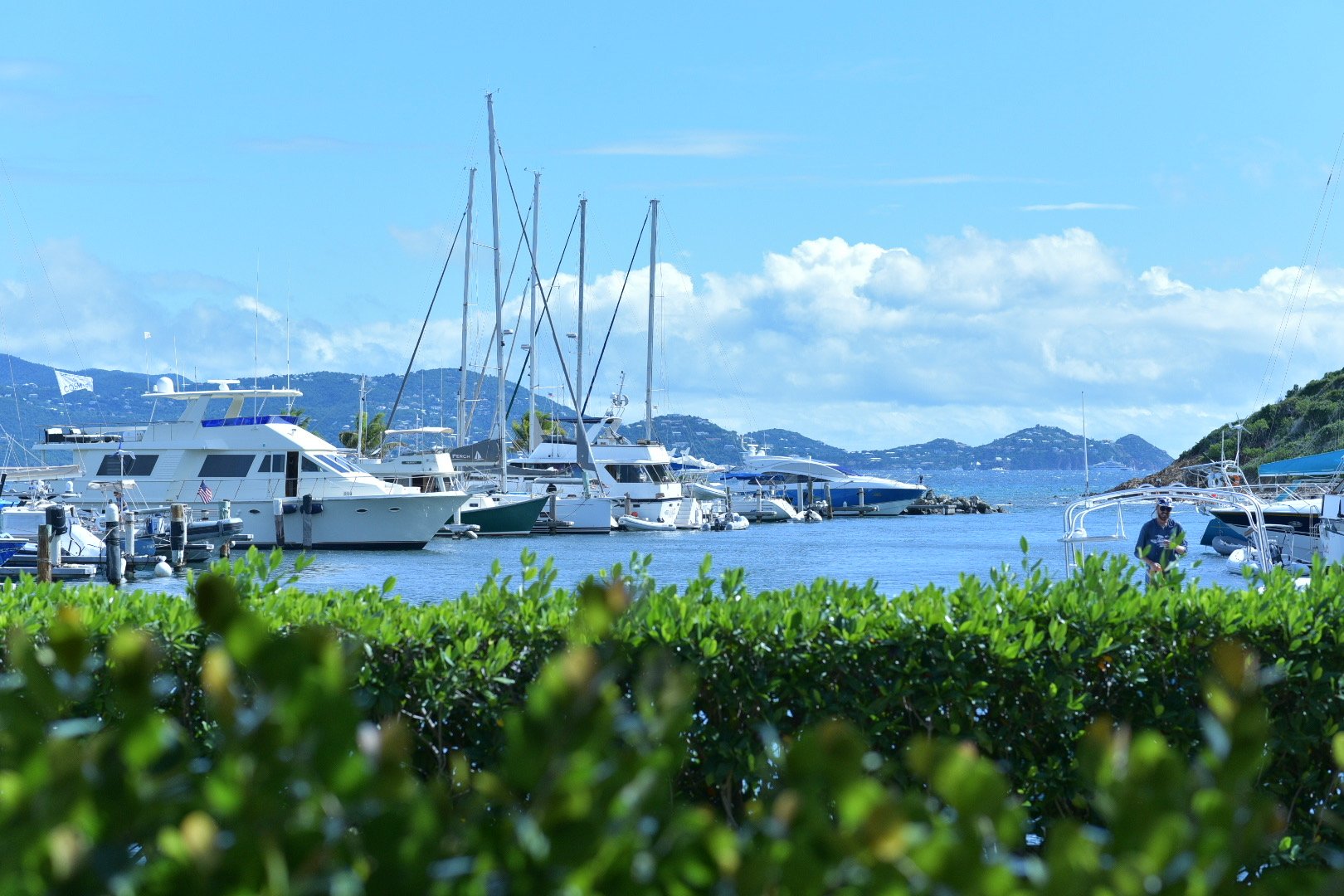 A marina with several boats and yachts docked, some with sails, against a backdrop of hills and a partly cloudy sky, with a person standing on the right near the water.