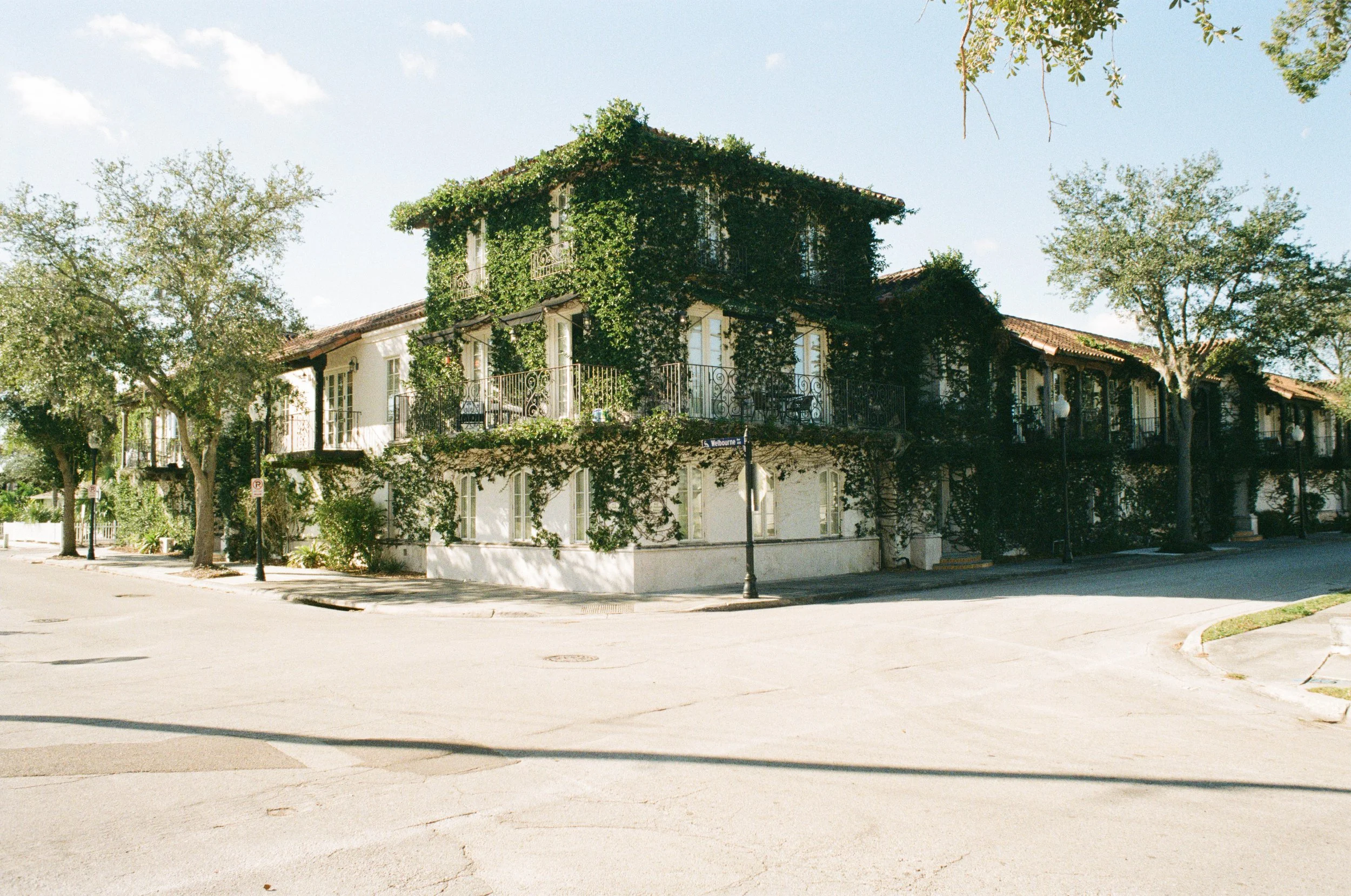 A three-story house with ivy growing on the exterior walls, featuring balconies with wrought iron railings, located on a quiet street corner with trees and street lamps, and a street sign reading 'Waldoore St' at the corner.