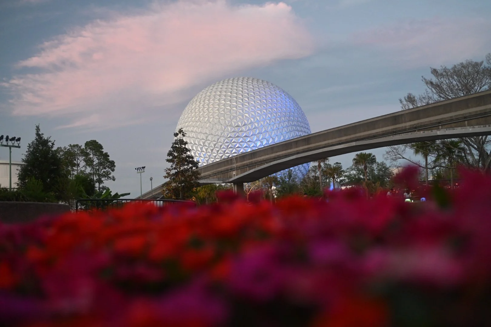 Epcot geodesic sphere at Disney World with a sky and trees, blurred pink flowers in the foreground.