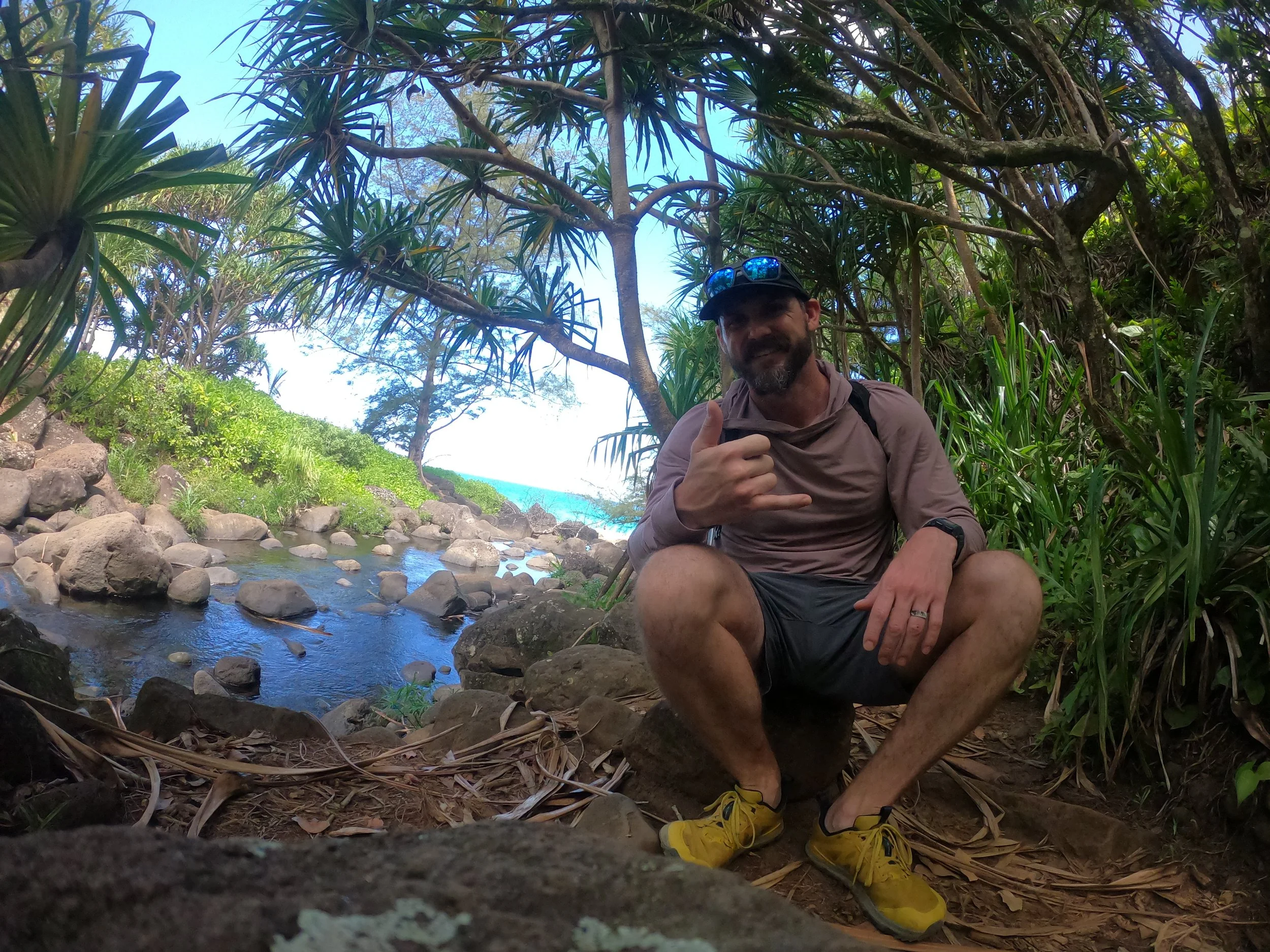 Man sitting on rocks near a small stream surrounded by lush green tropical plants and trees, with a view of the beach and ocean in the background, giving a thumbs-up gesture and smiling, wearing sunglasses on his cap and yellow hiking shoes.