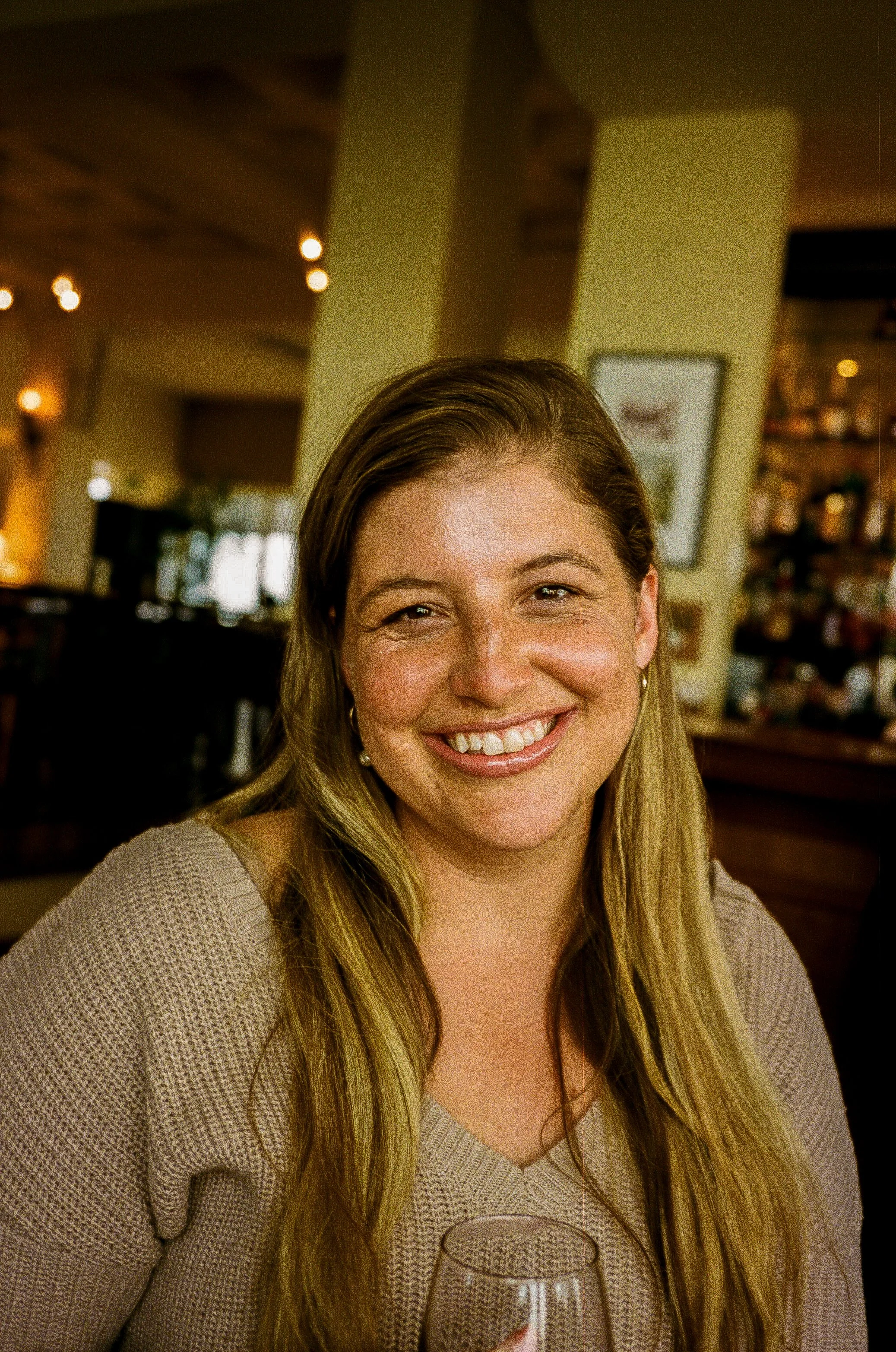 A smiling woman with long blond hair, wearing a beige knit sweater, sitting in a restaurant or cafe, holding a wine glass, with warm lighting and blurred background.